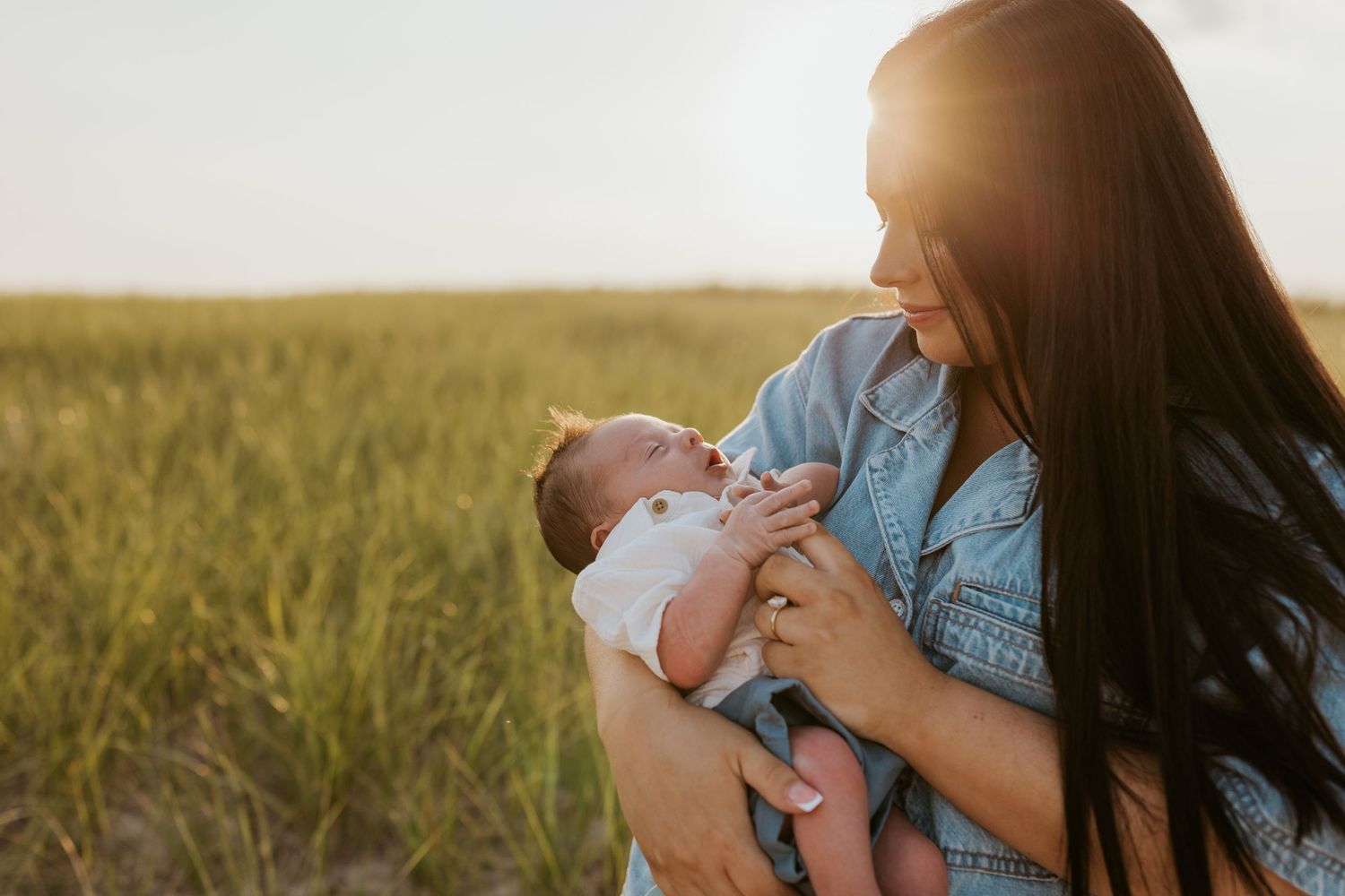 Mother in denim shirt holding newborn baby in golden sunset light in lush green field.