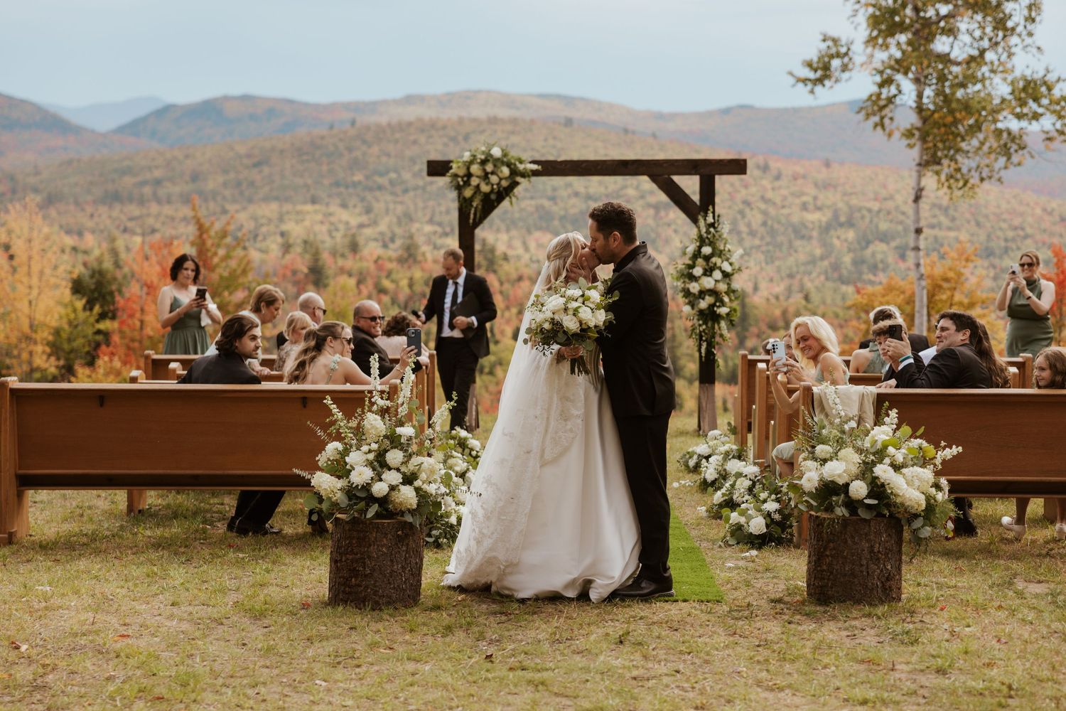 Outdoor fall wedding ceremony with couple kissing at wooden arch, floral arrangements, mountain backdrop, and seated guests.