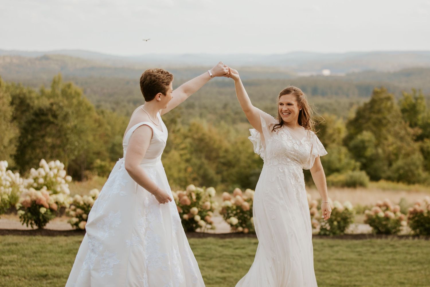 Two brides in white wedding gowns dancing and twirling together in a scenic garden landscape.