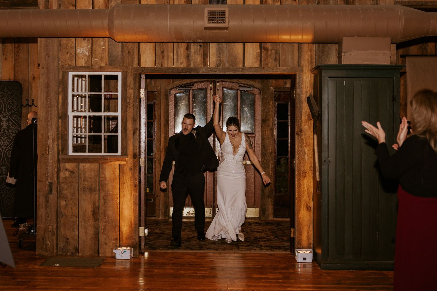 Bride and groom make grand entrance at rustic barn wedding reception, raising hands in celebration on hardwood dance floor.