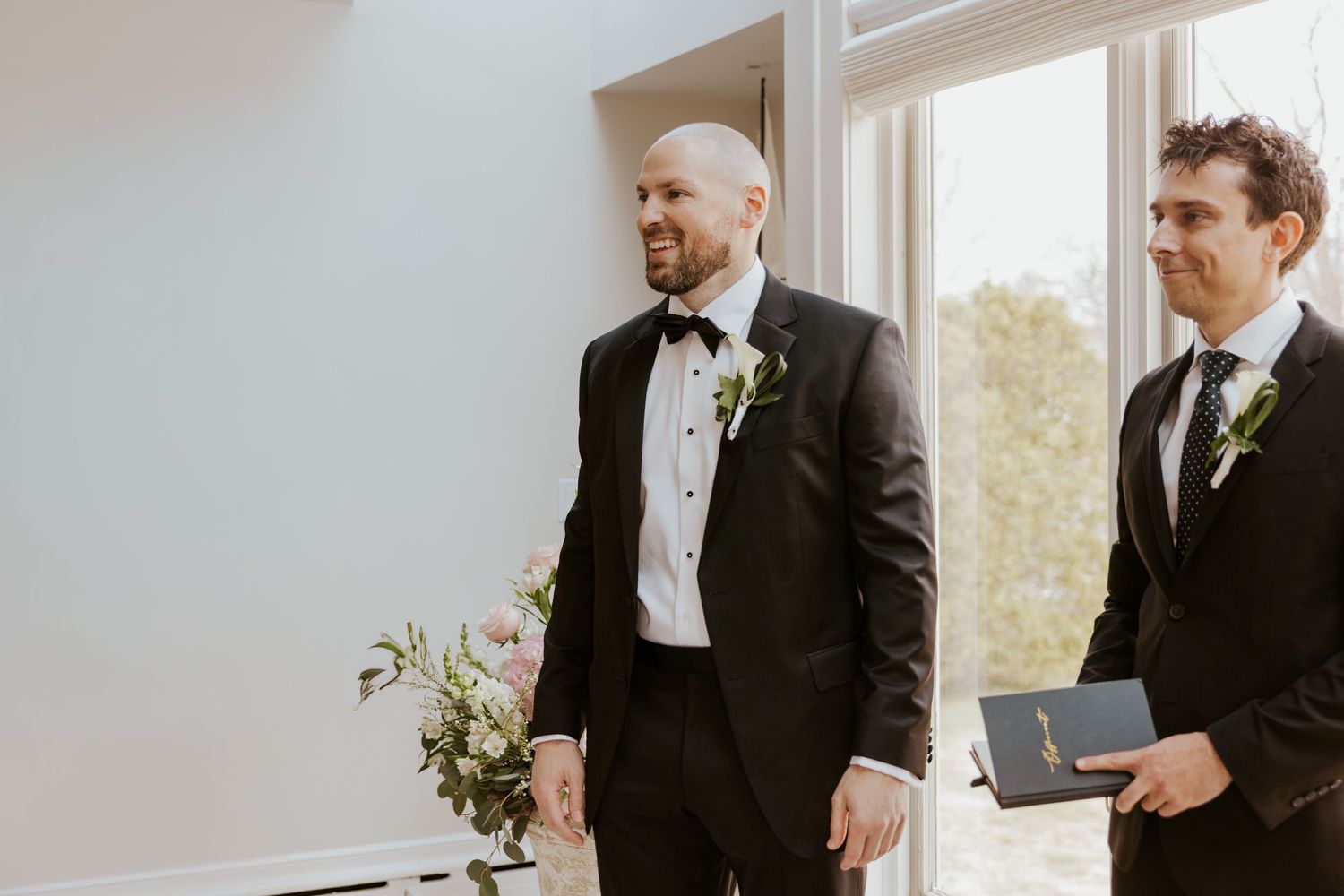 Groomsmen share a lighthearted moment during formal wedding preparations in a bright indoor setting.