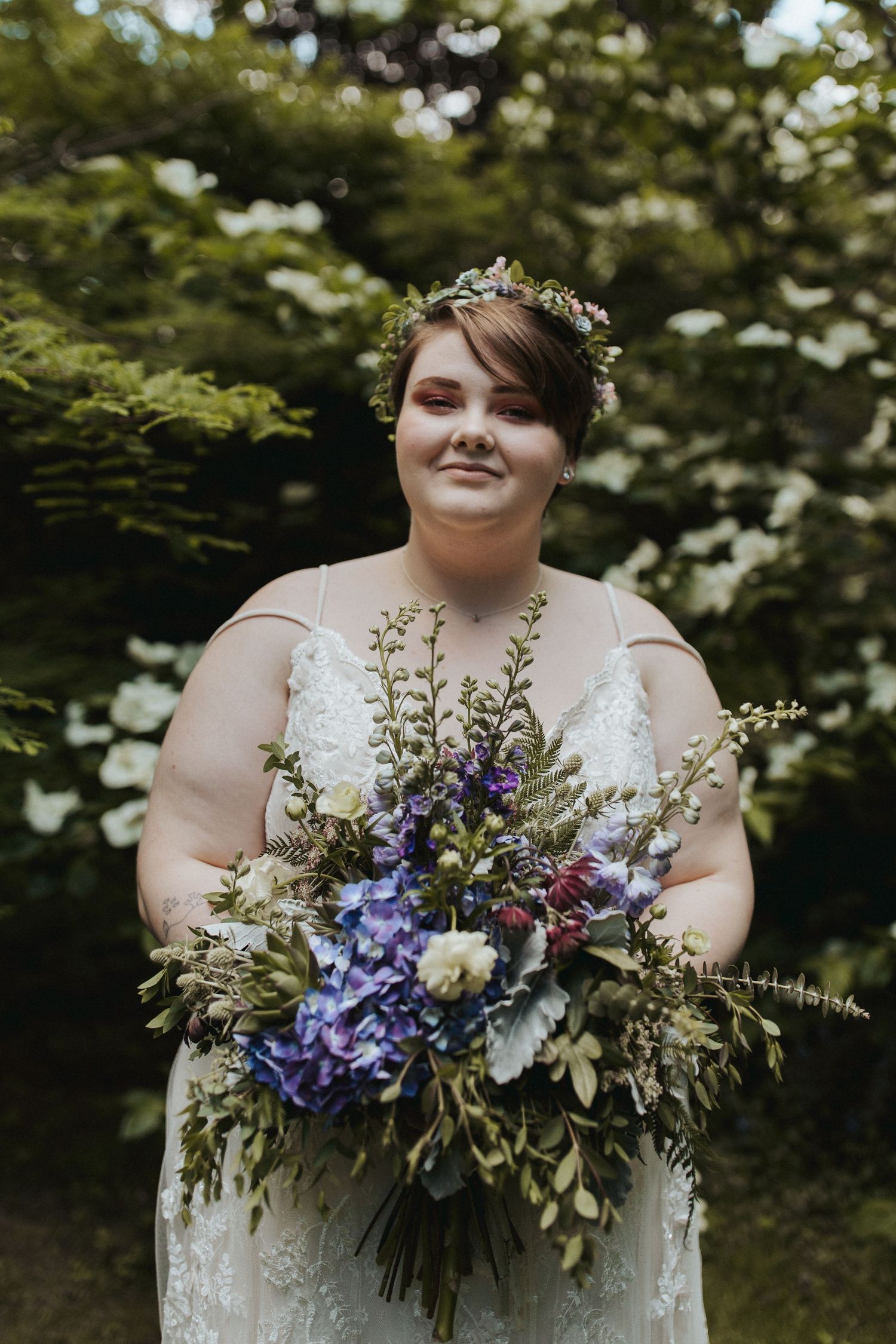 A person in a white wedding dress holds a stunning purple and blue floral bouquet outdoors.