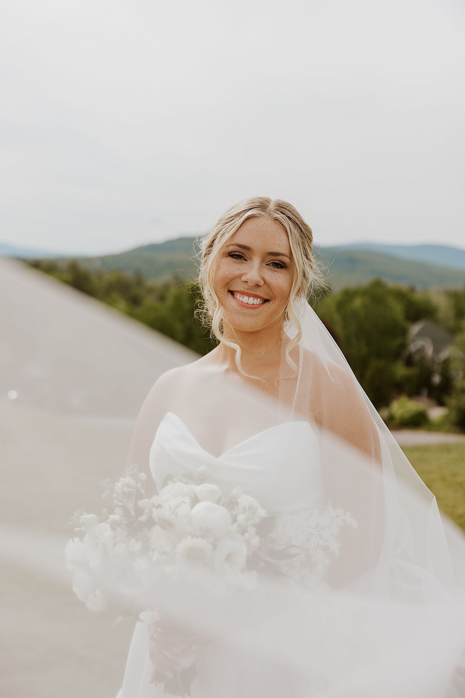 Smiling bride in white gown holds bouquet, veil flowing in breeze, with green hills backdrop.