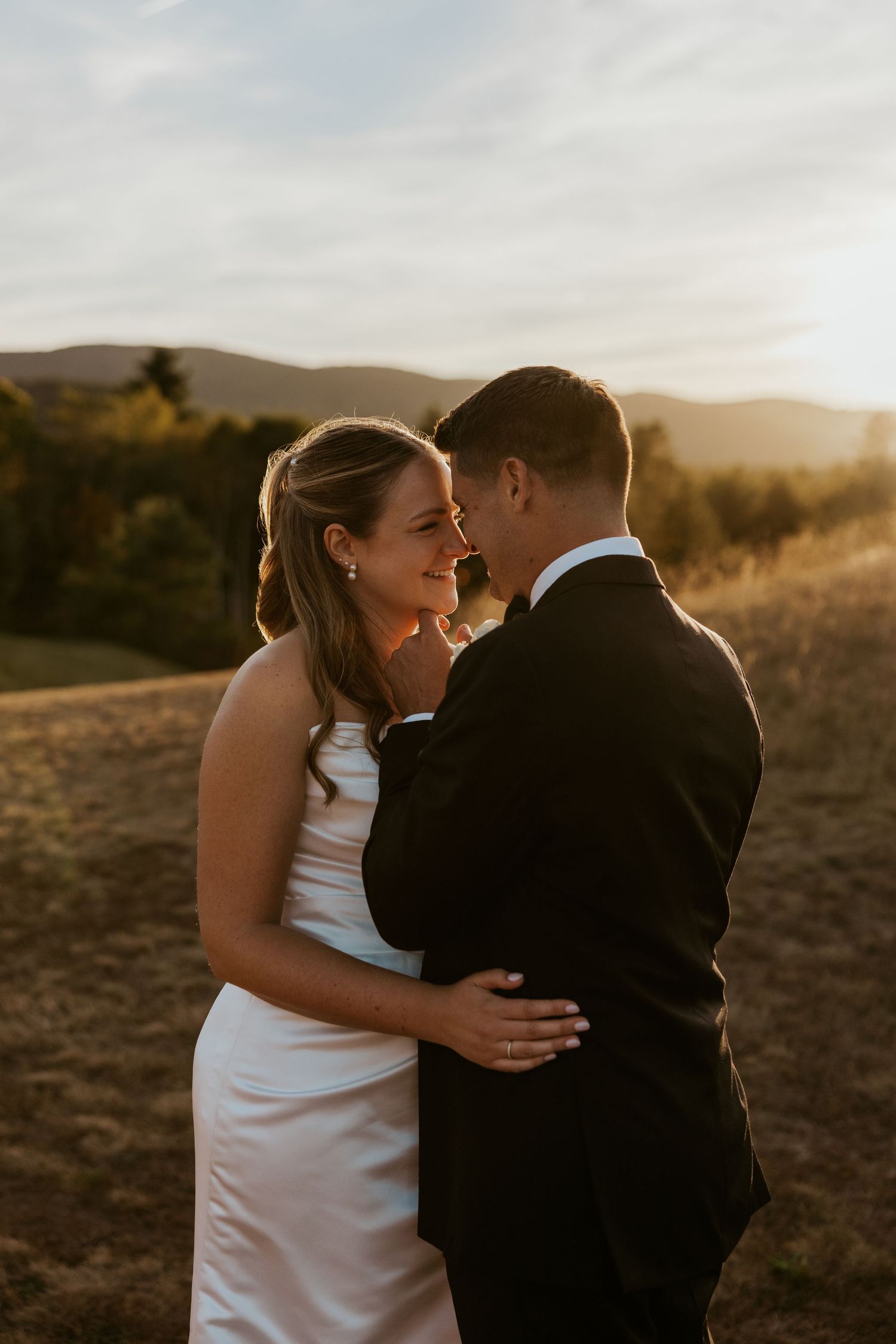 Bride in white gown and groom in black suit embrace at sunset with golden hills and mountains in the background.