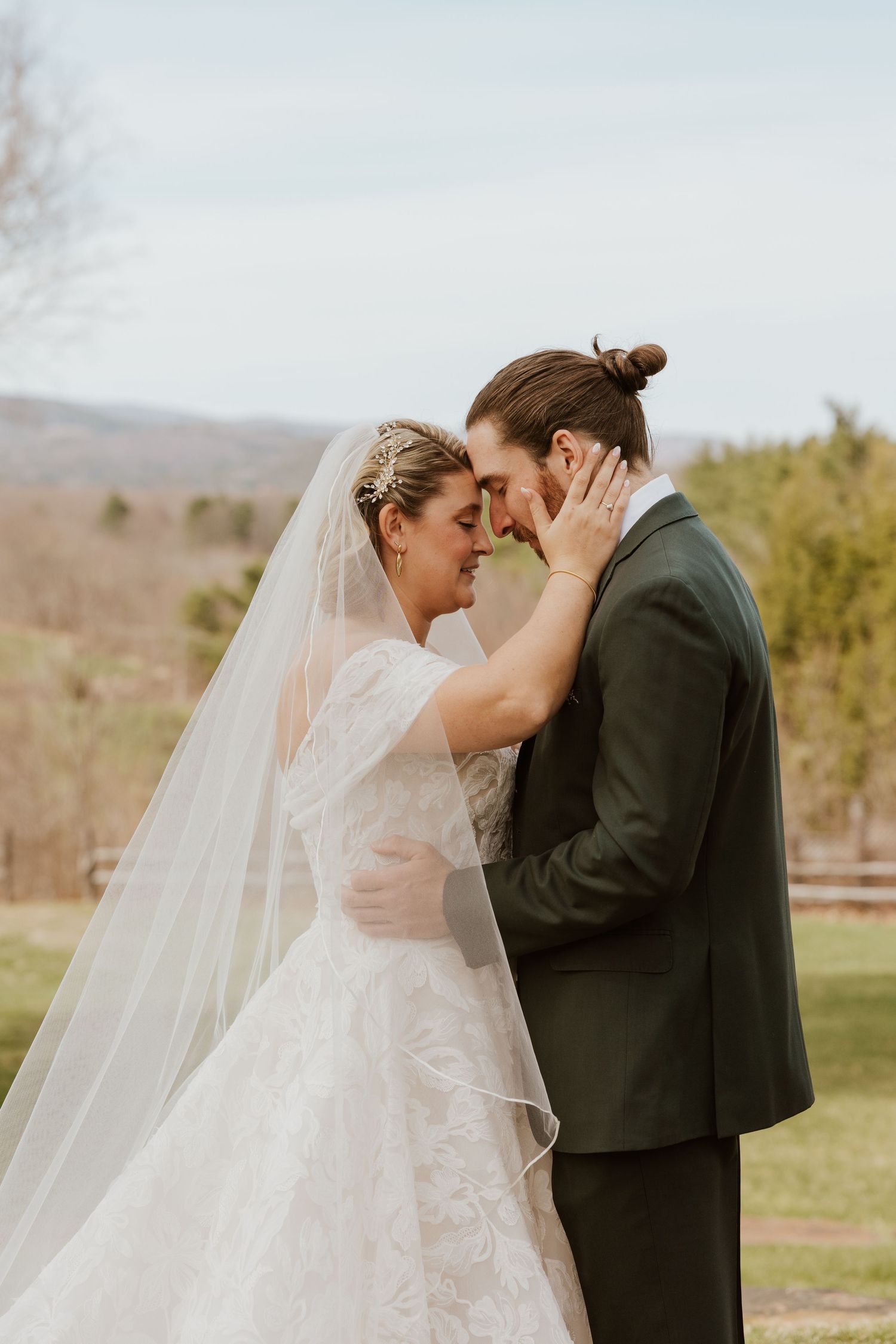 Bride and groom touching foreheads tenderly in an outdoor wedding portrait with scenic hills behind.