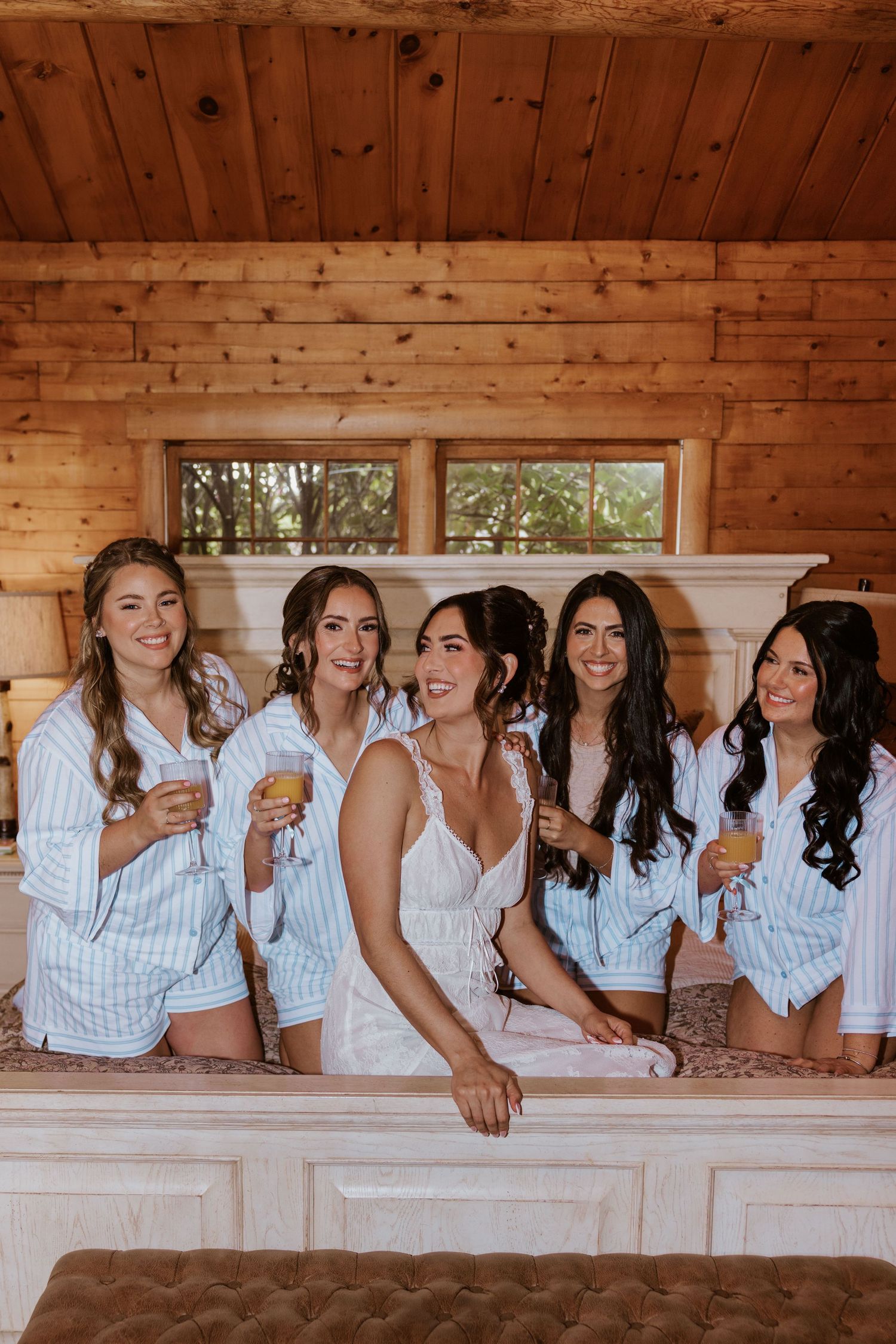 Bride and bridesmaids in matching striped pajamas smiling together in a rustic wood-paneled room during wedding preparations.