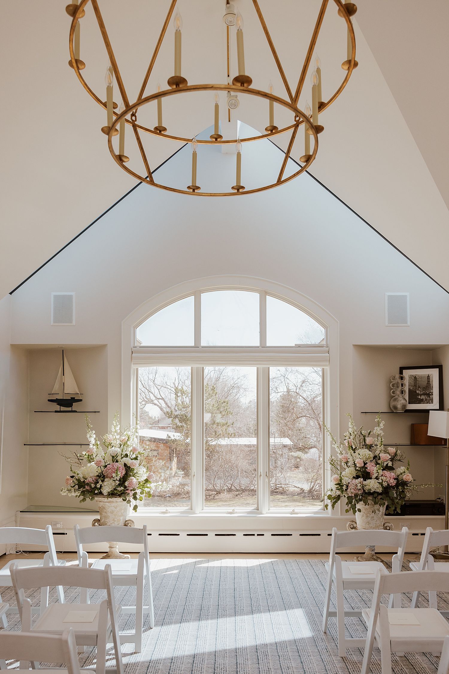 Elegant wedding chapel with white chairs, floral arrangements, arched window, and gold chandelier under vaulted ceiling.
