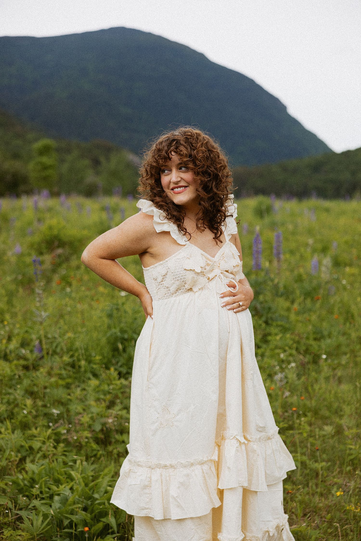 Smiling pregnant woman in cream ruffled dress posing in wildflower meadow in New Hampshire taking motherhood photos