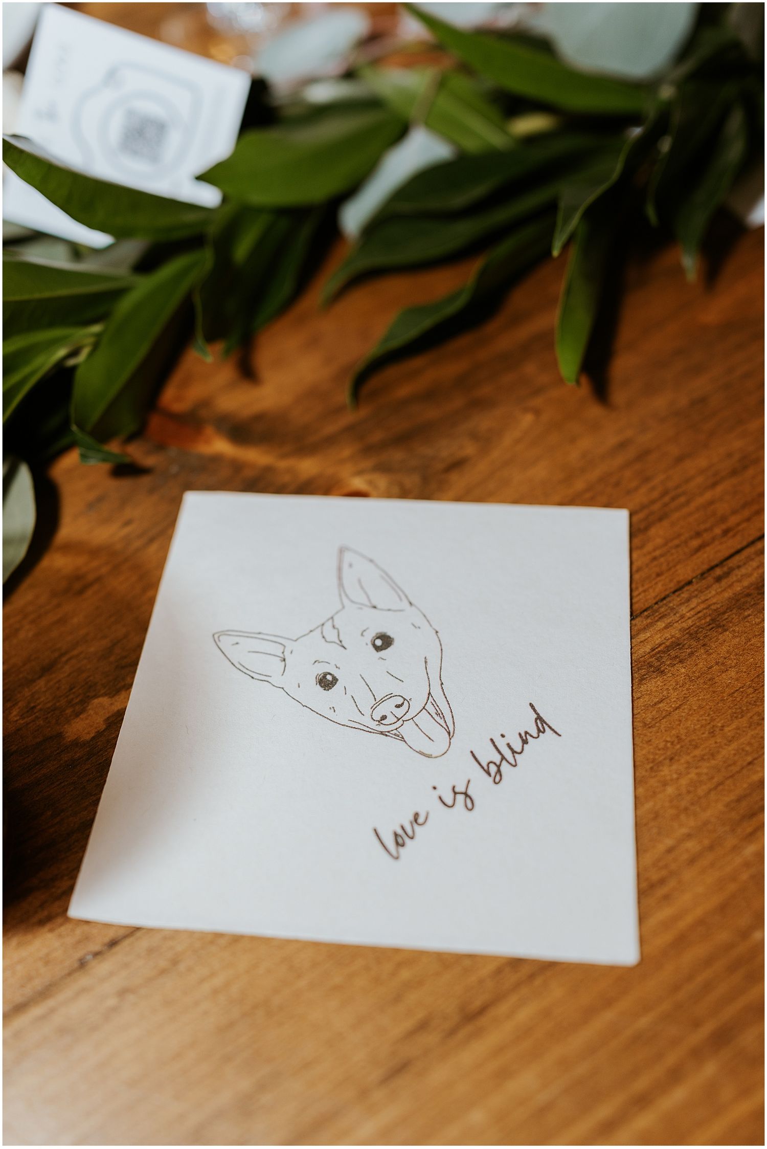 Simple hand-drawn cat illustration with 'love u' text on white paper against wooden surface with green leaves.