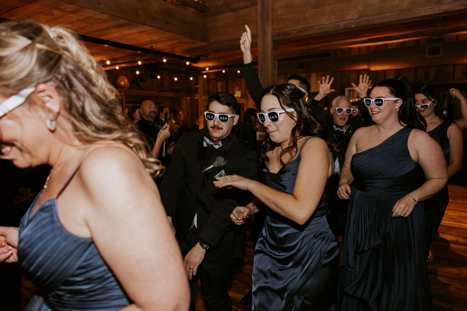 Wedding guests dancing joyfully in sunglasses at a lively indoor reception party.