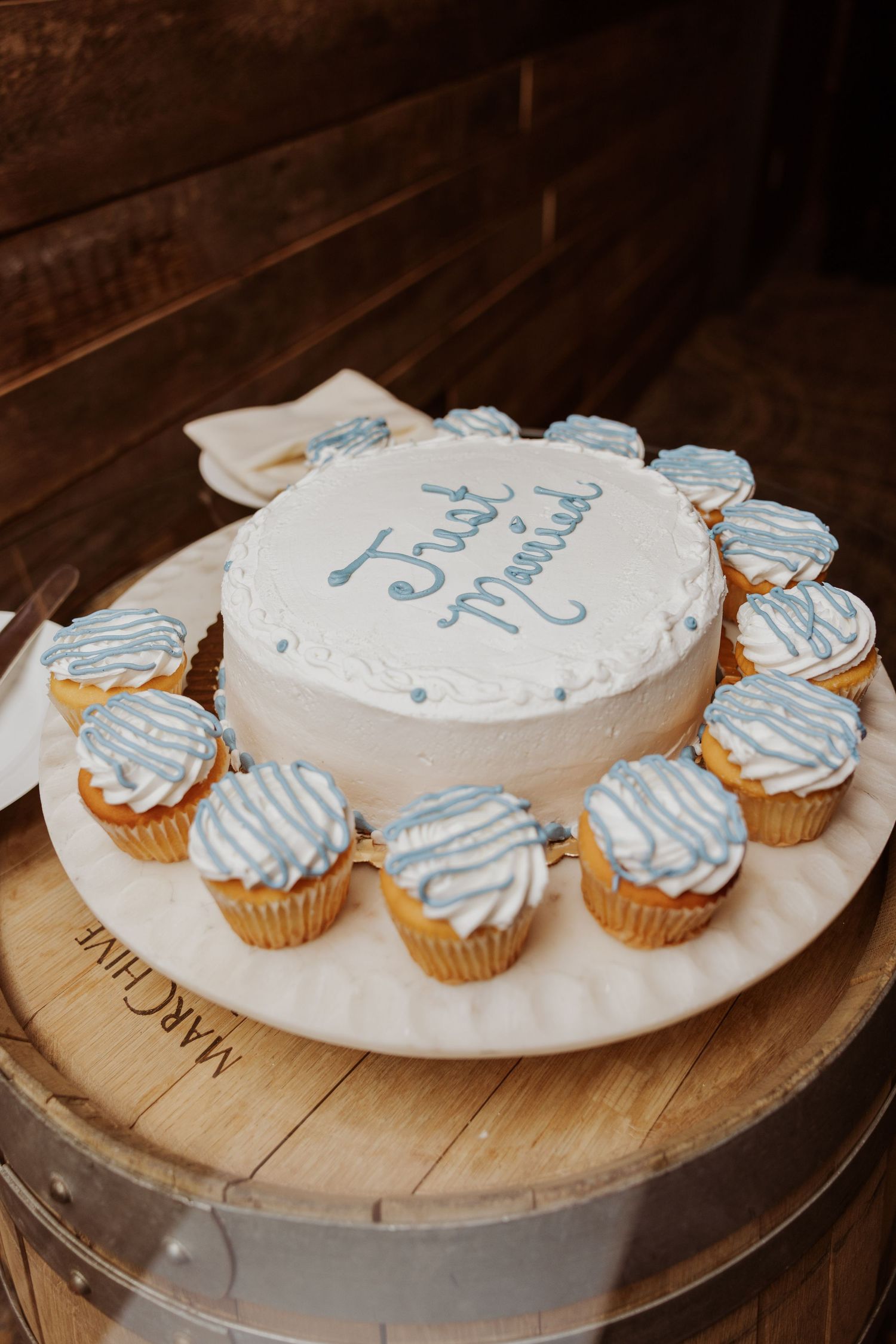 A white frosted cake with blue striped cupcakes arranged around it sits on a wooden serving board.