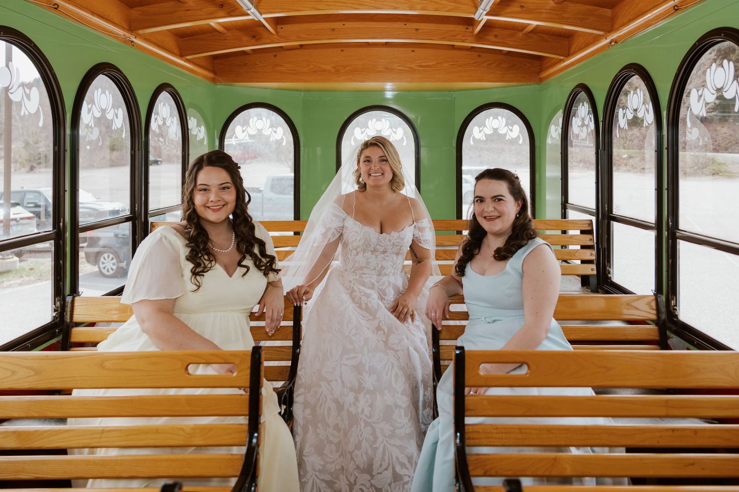 Bride in lace gown poses with two bridesmaids inside a vintage green trolley on her wedding day.