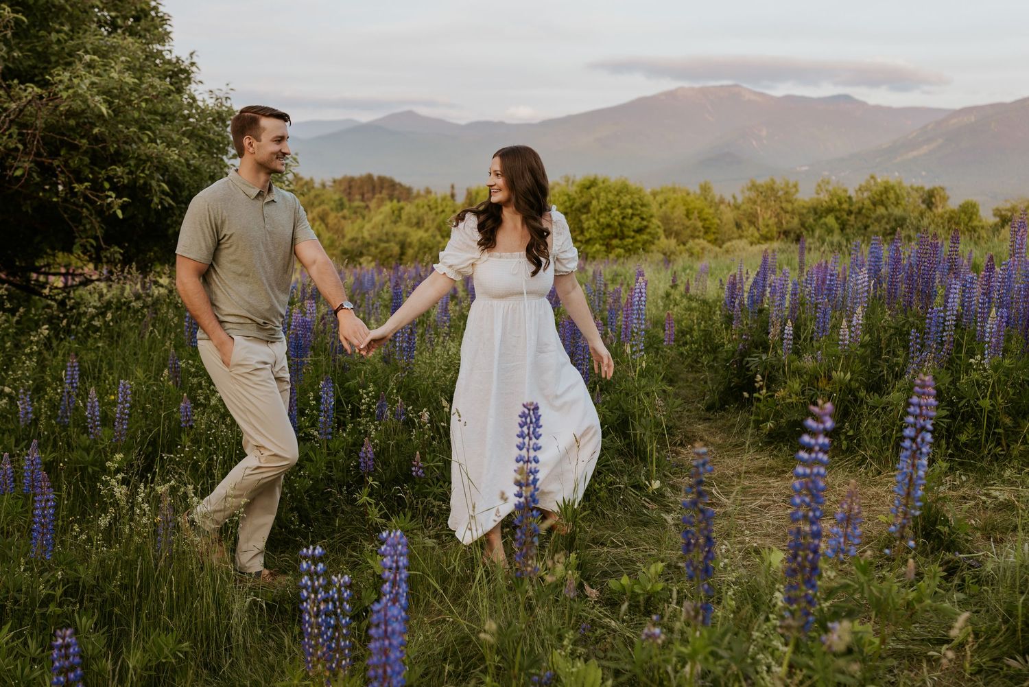 Happy couple walking hand in hand through a vibrant purple lupine field at sunset.