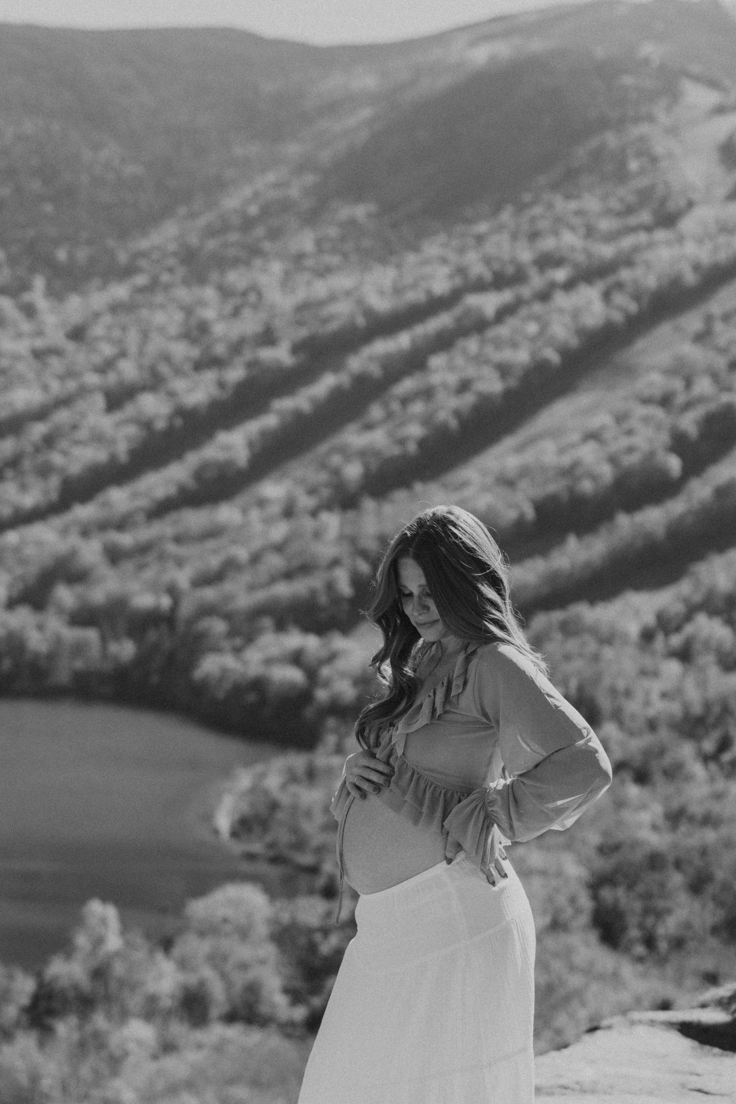Pregnant woman in white skirt poses on mountain overlook with scenic valley and forest backdrop.