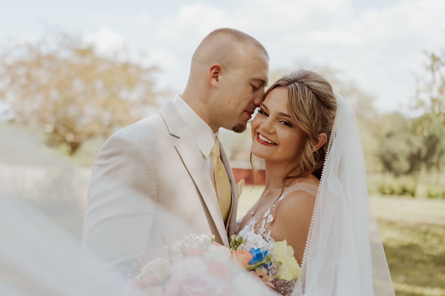 Groom tenderly kisses bride on the forehead as she smiles holding her wedding bouquet outdoors.