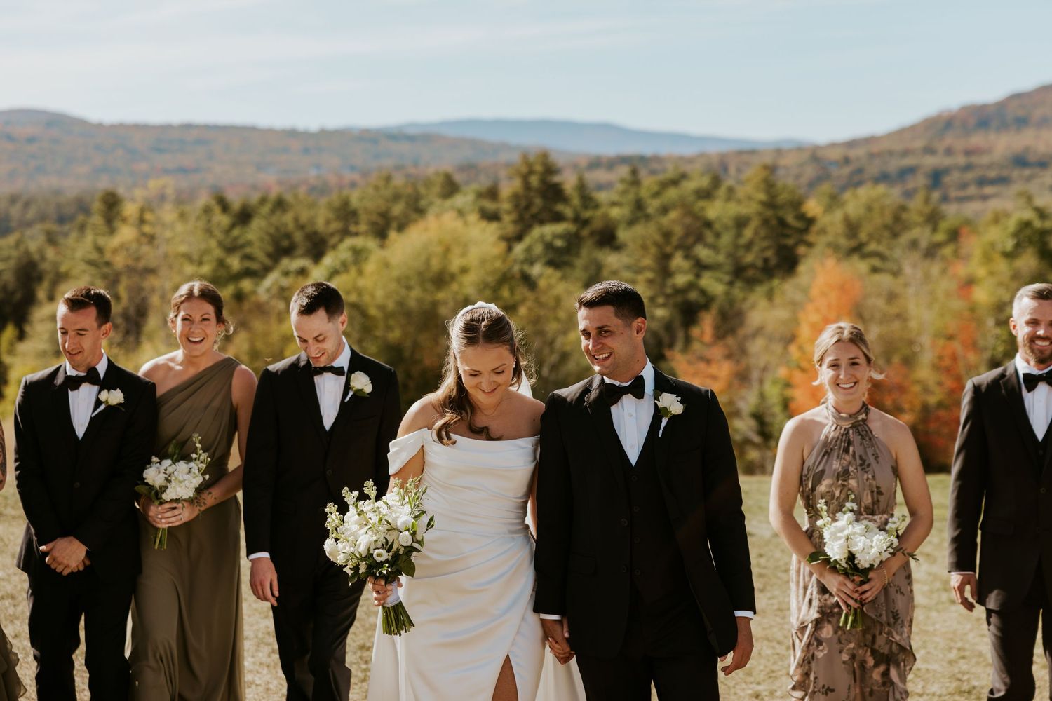 Wedding party walking outdoors in fall foliage with bride in white gown and groomsmen in black tuxedos.