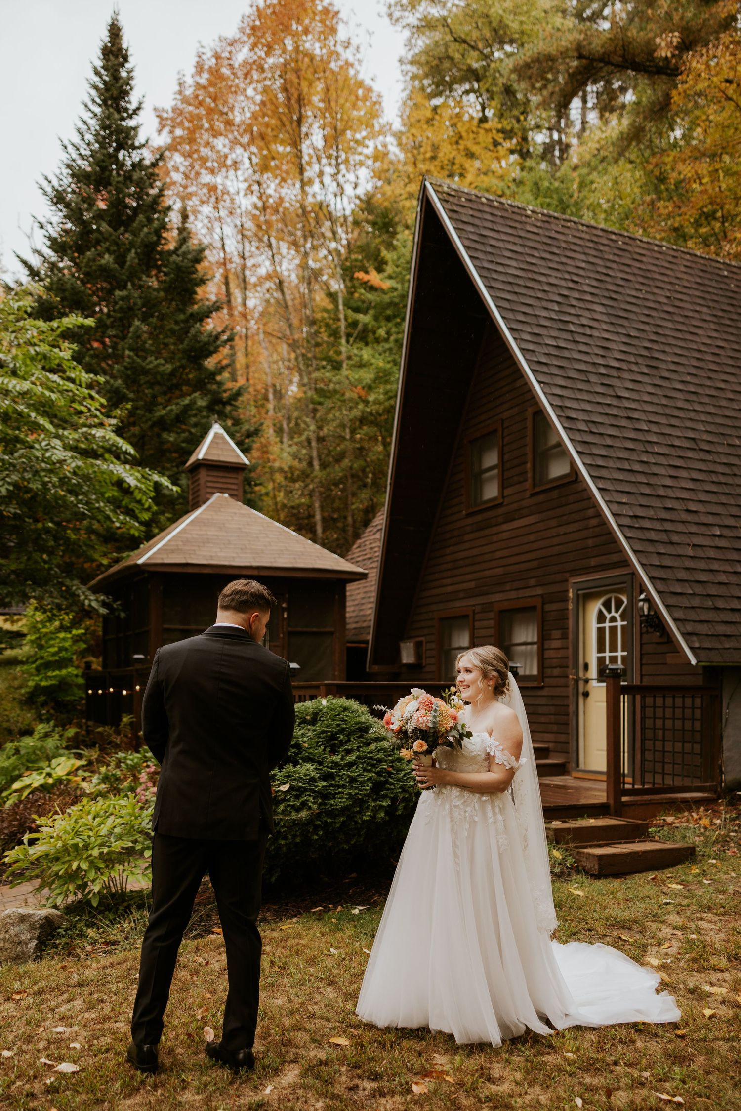 Bride in lace ball gown holds bouquet during first look moment outside rustic A-frame cabin in fall.