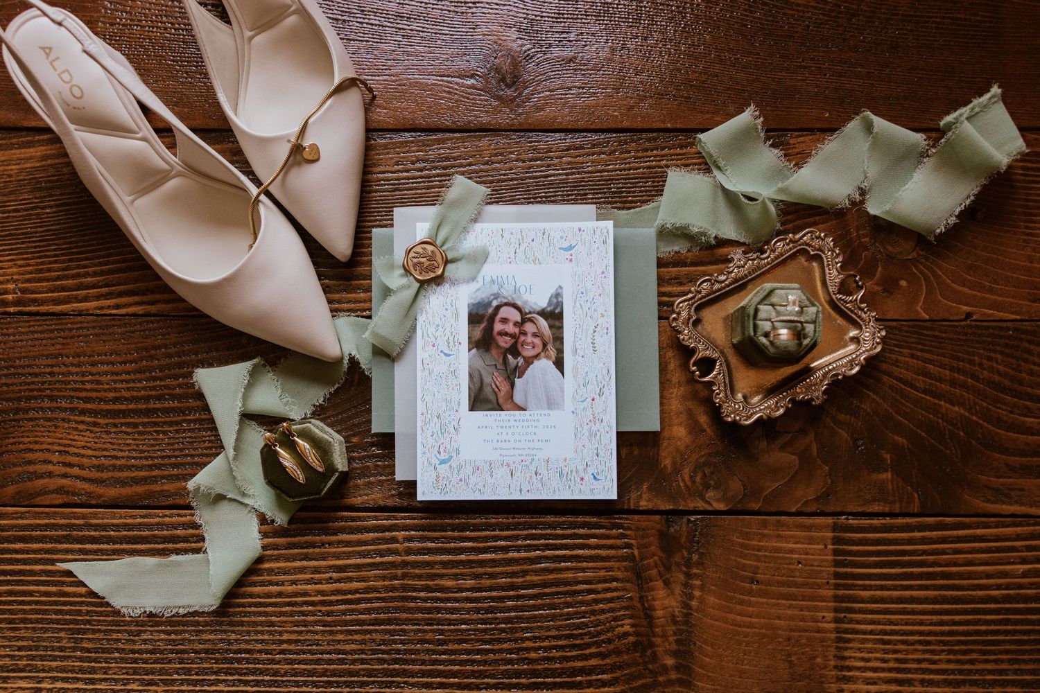 Wedding flat lay featuring nude heels, sage green ribbon, photo card, and gold ring dish on rustic wood.