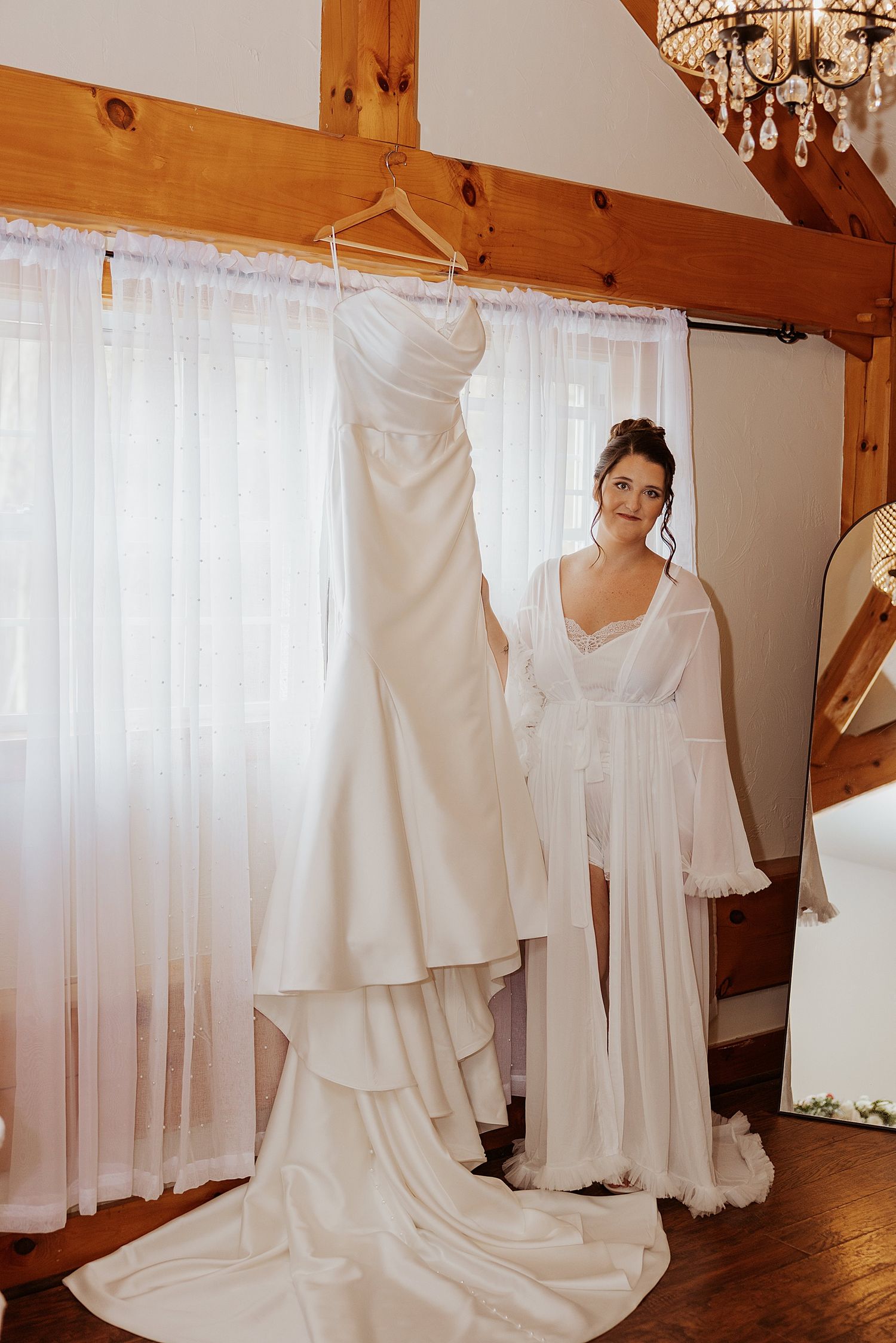 Bride in white robe holding her elegant strapless wedding gown on a hanger in a rustic bridal suite in NH