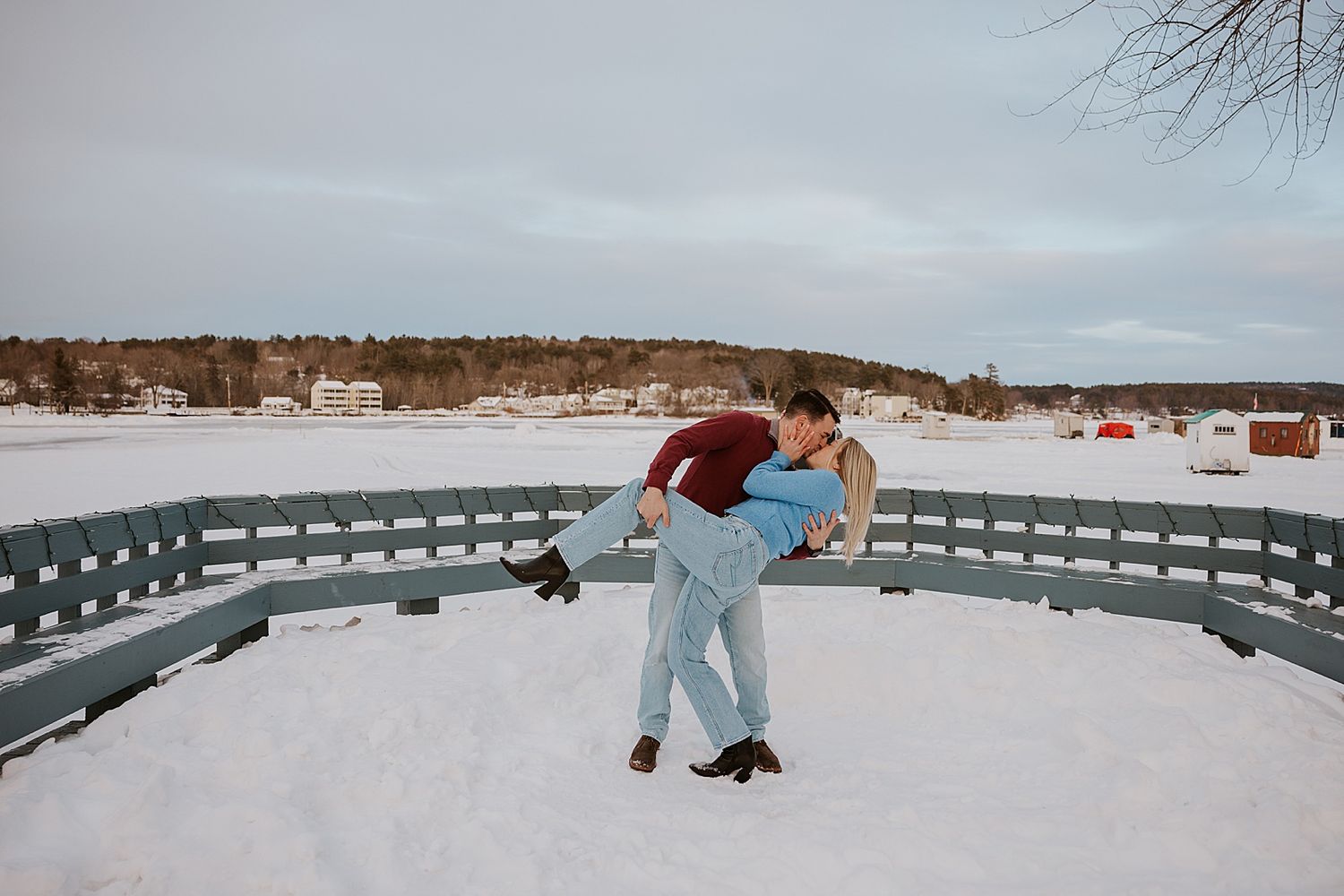 Couple sharing a romantic dip pose in snowy winter landscape near a curved bench outdoors.