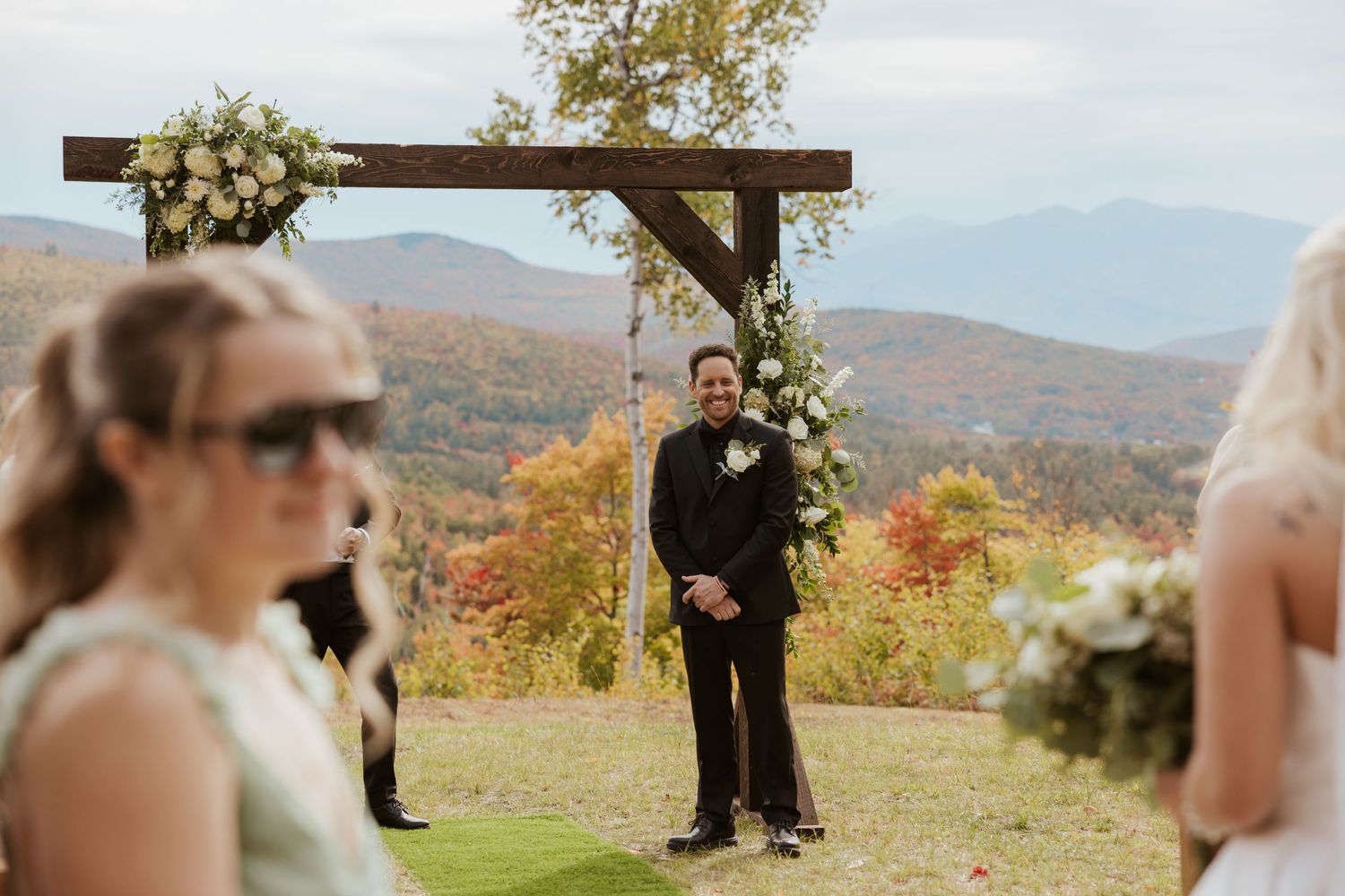 Groom smiling at altar with floral arch and stunning mountain fall foliage backdrop at outdoor wedding ceremony.
