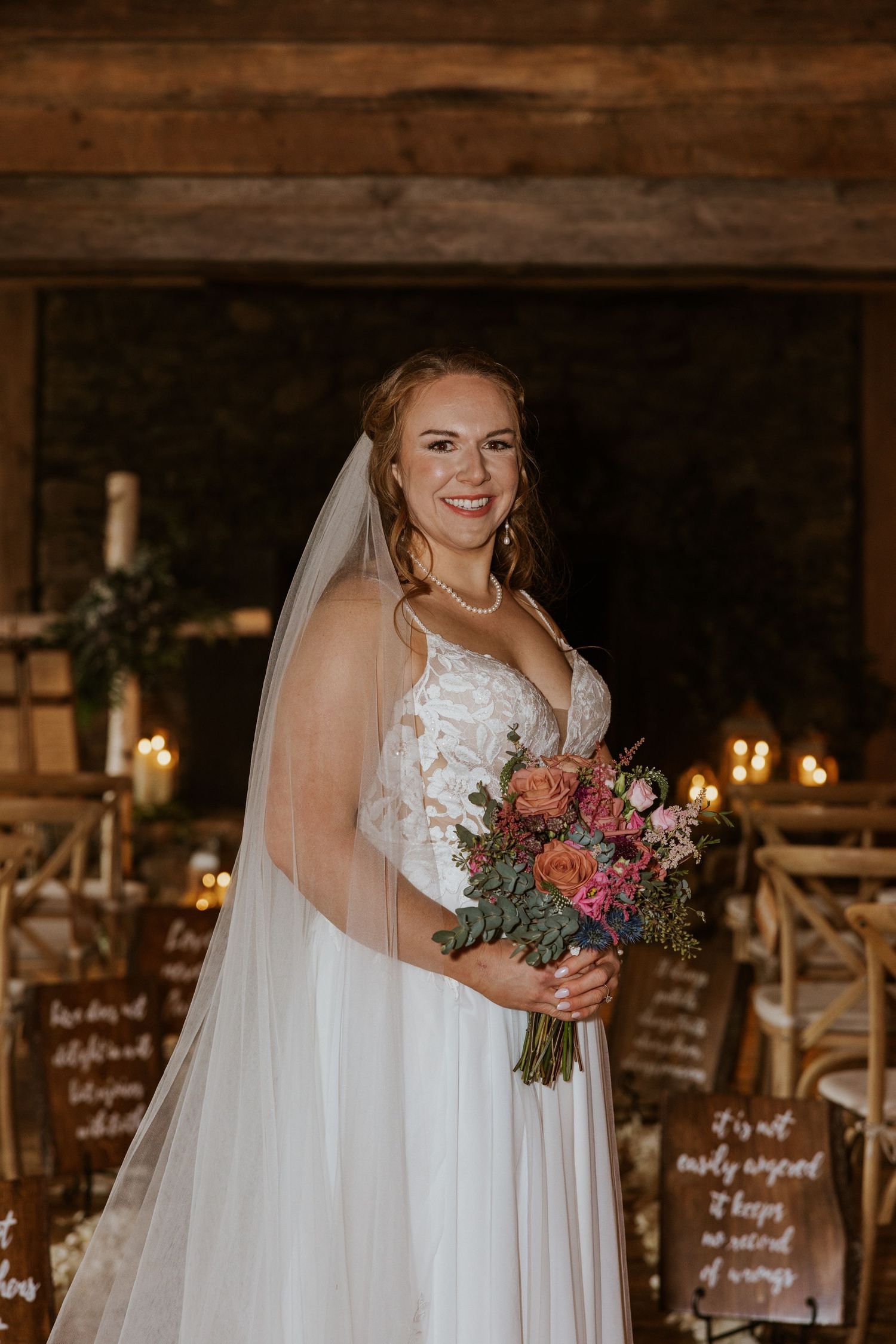 Bride in white lace wedding dress holds colorful bouquet in rustic barn setting with candlelight.