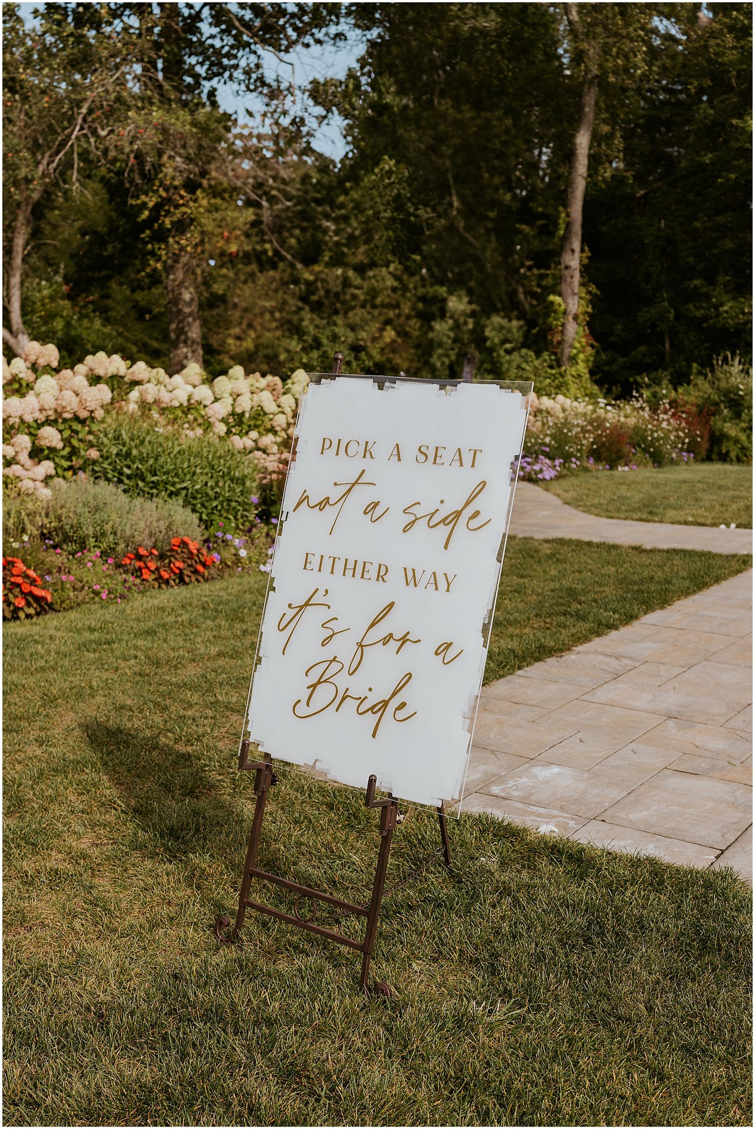 Elegant wedding ceremony signs in white with gold calligraphy displayed on a manicured lawn direct guests to seating.
