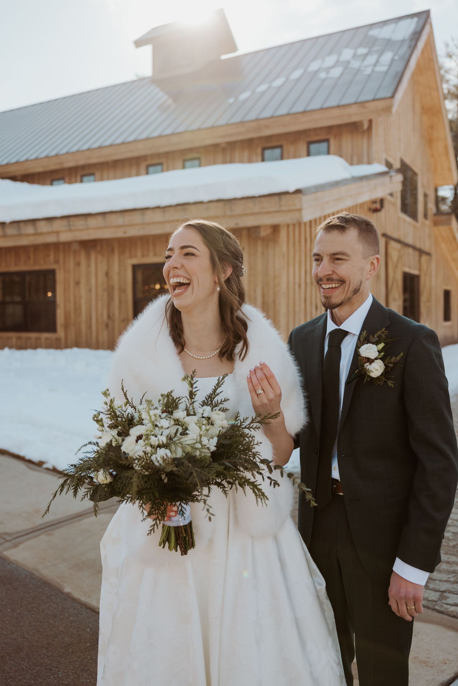A joyful wedding couple shares candid laughs together in a rustic barn setting during their winter celebration.