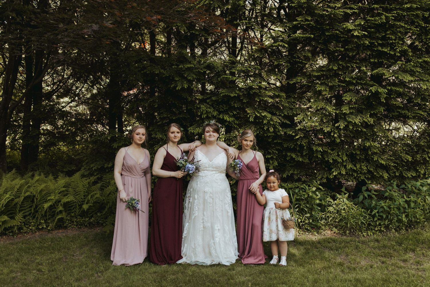 Wedding party in pink and burgundy dresses pose together outdoors.