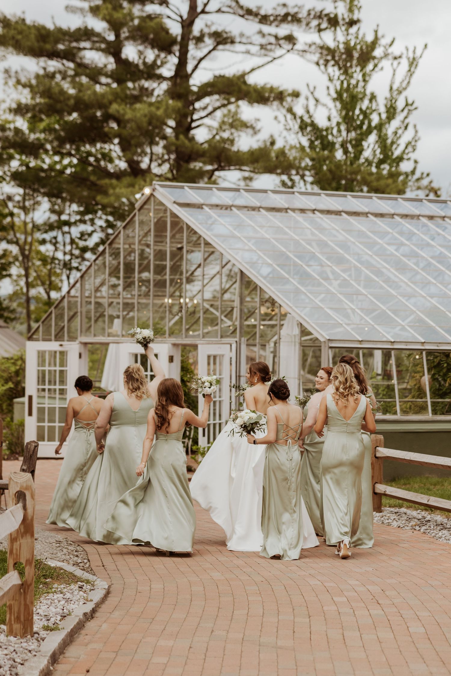 Bride walks with bridesmaids in sage green gowns toward a glass greenhouse wedding venue.
