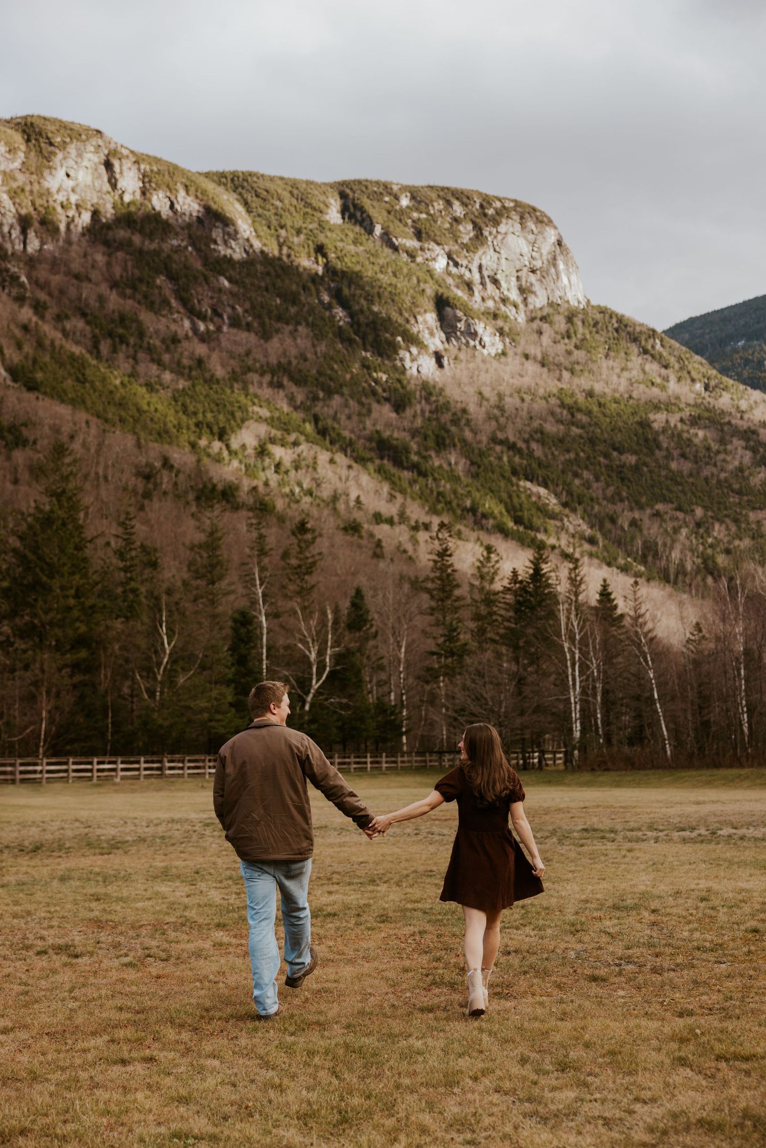 Couple strolling hand in hand through an open field with a dramatic rocky mountain and pine forest behind them.