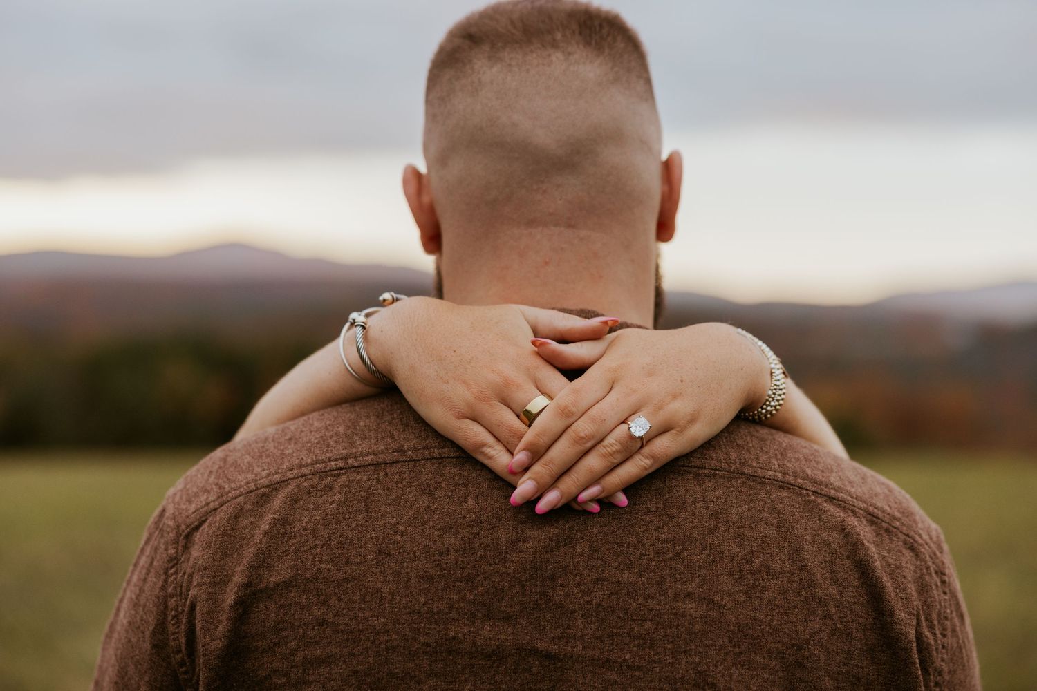 Close-up of womans hands with engagement ring wrapped around mans shoulders in an autumn outdoor setting.