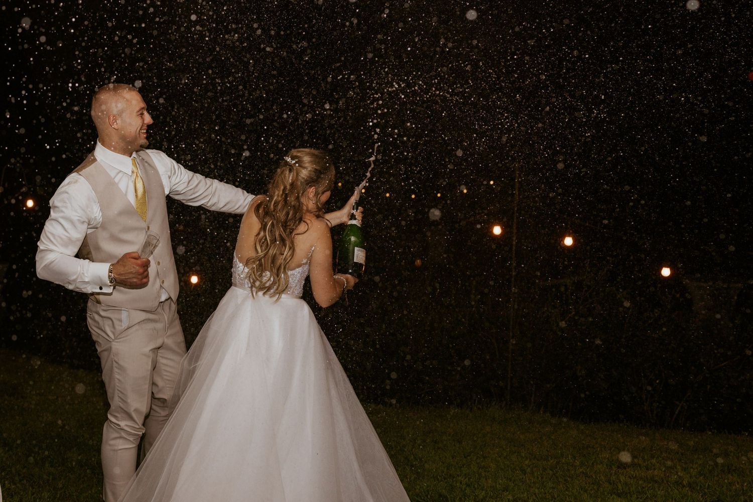 Bride and groom celebrating with champagne spray under romantic string lights at night.