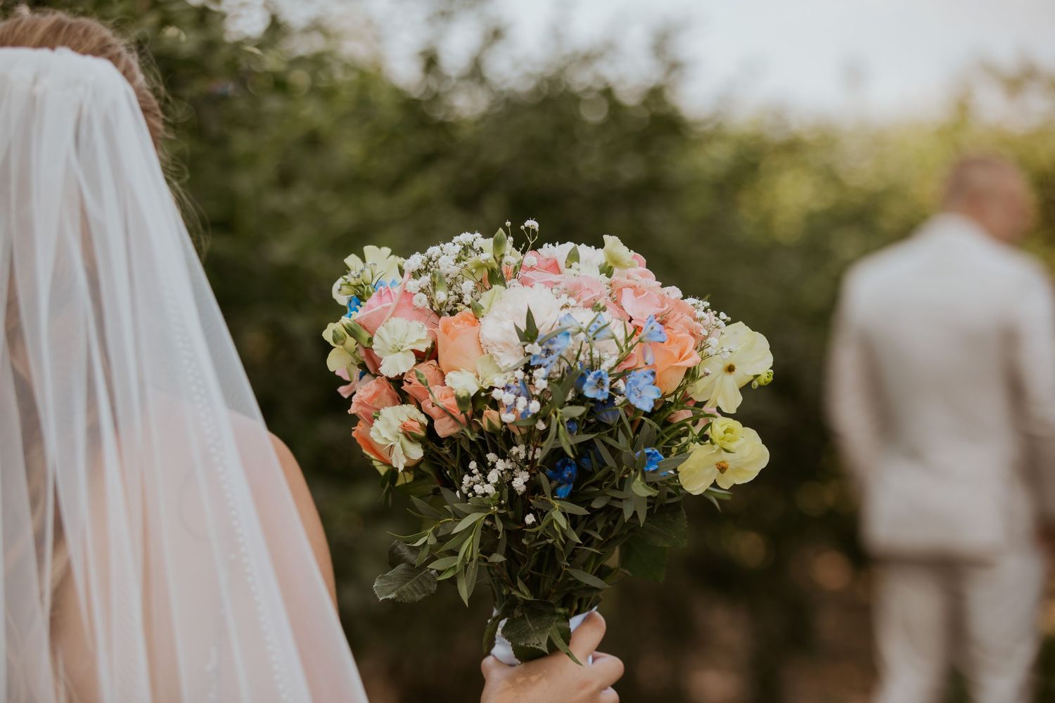 Bride holding a vibrant colorful wedding bouquet with pink, blue, and yellow flowers with greenery.