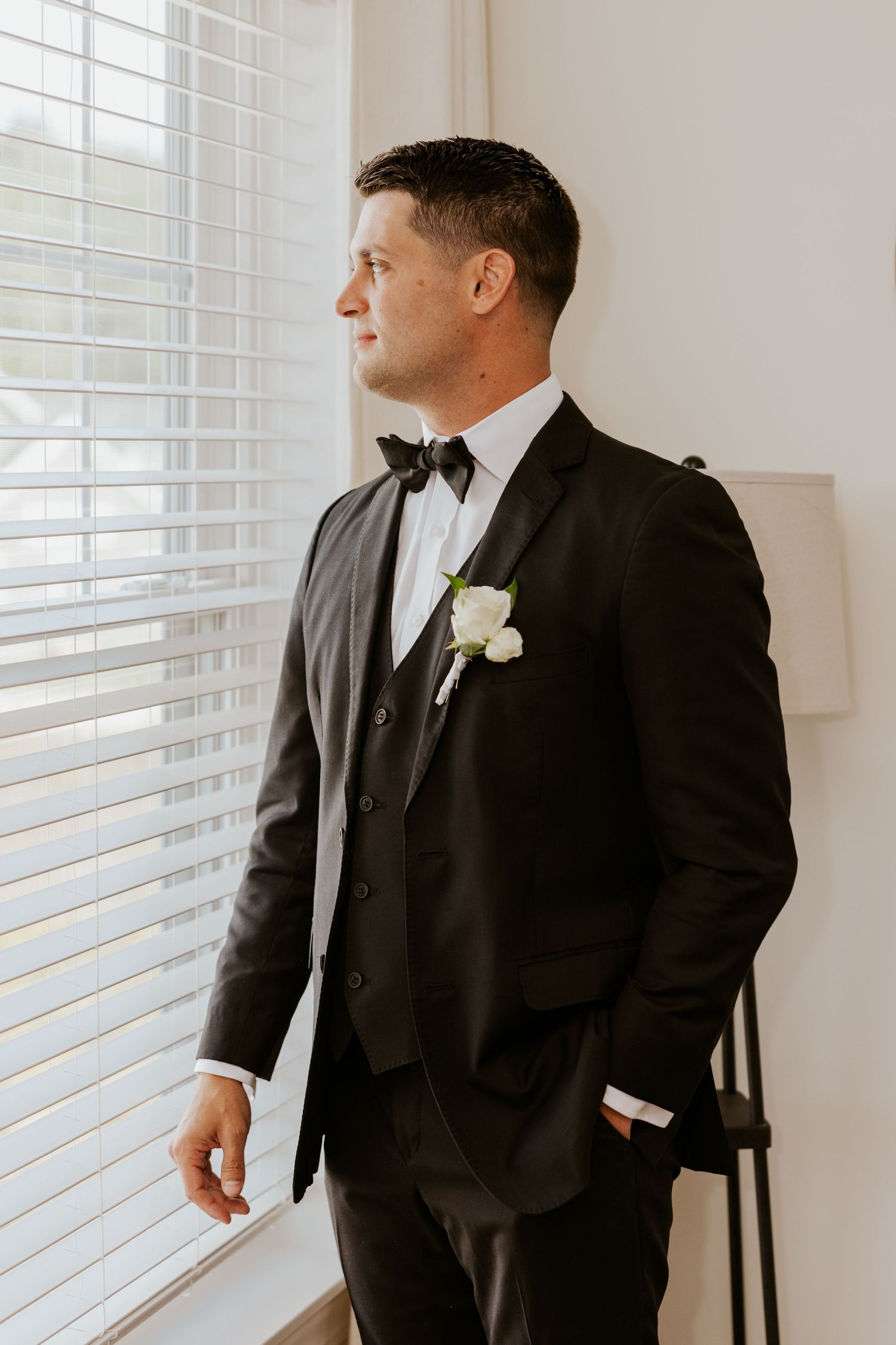 Handsome groom in black tuxedo with bow tie and white boutonniere gazing out a bright window.