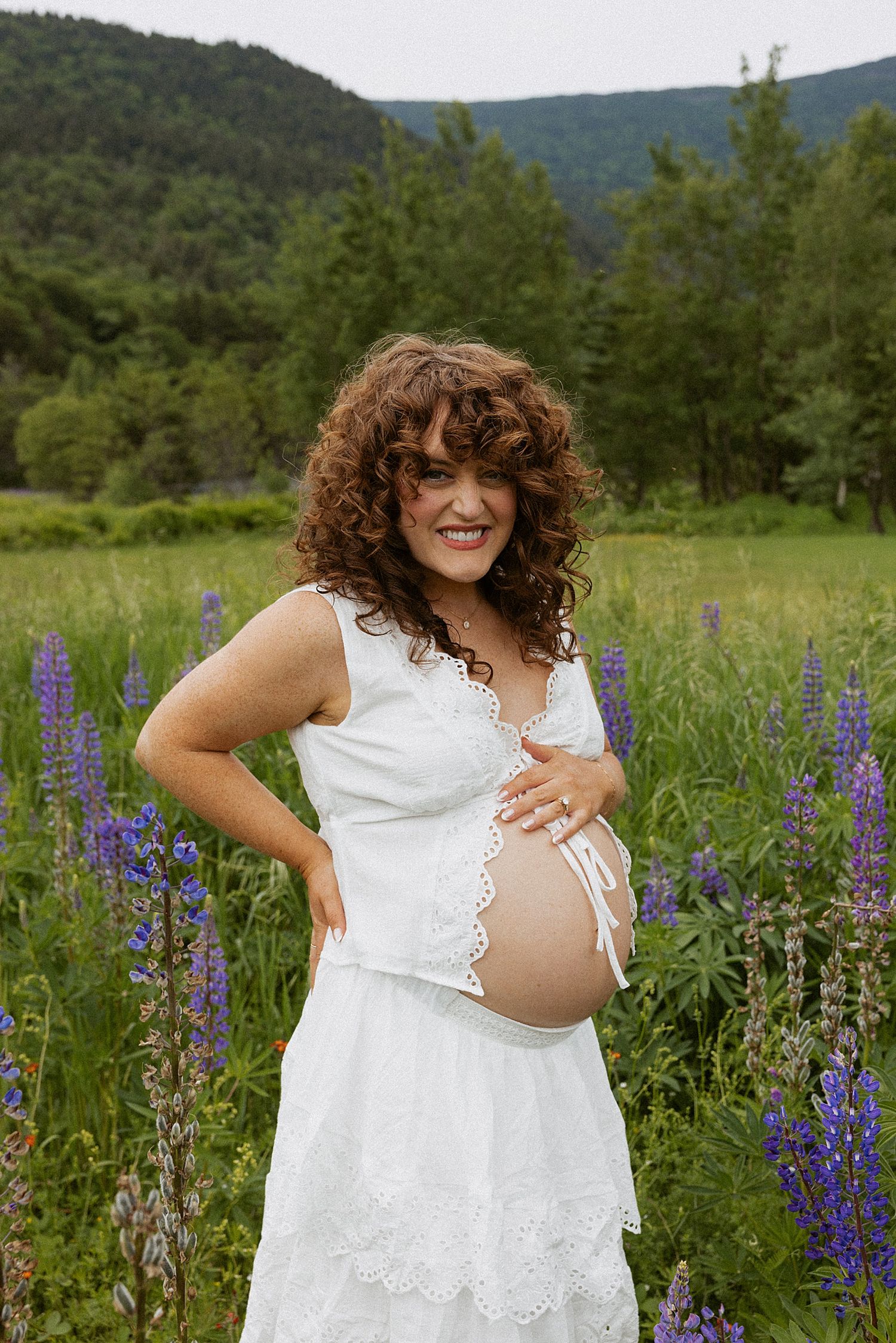 Smiling pregnant woman in white dress holding baby bump in a field of purple lupine flowers with mountains behind in New Hampshire