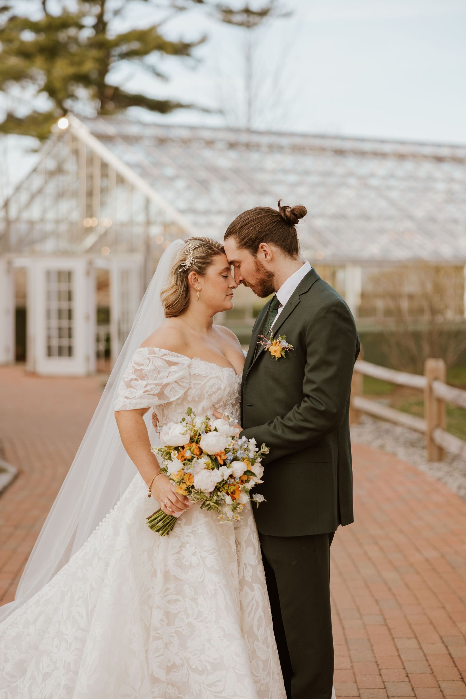 Bride and groom touching foreheads tenderly, she holds a white bouquet, standing before a greenhouse backdrop.