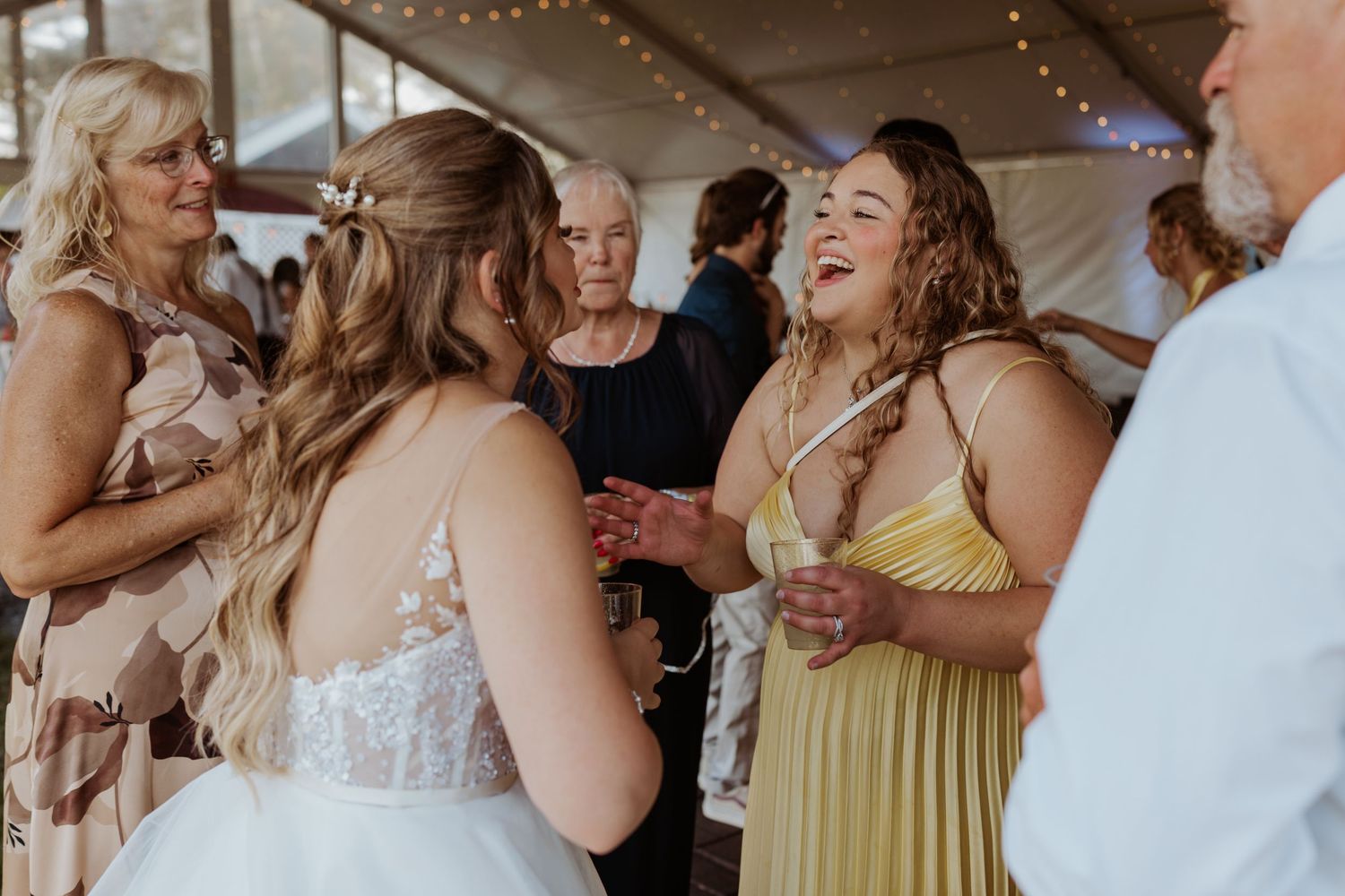 Bride laughing with bridesmaid in yellow dress at outdoor wedding reception under tent with string lights.