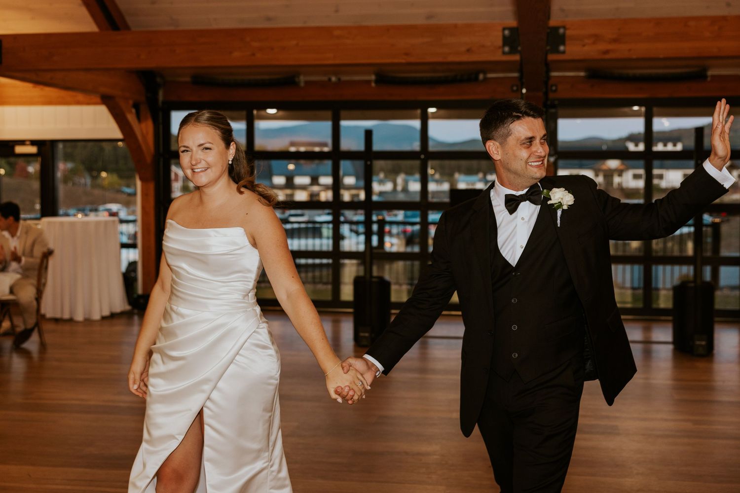 Smiling bride and groom hold hands while walking across dance floor at wedding reception.