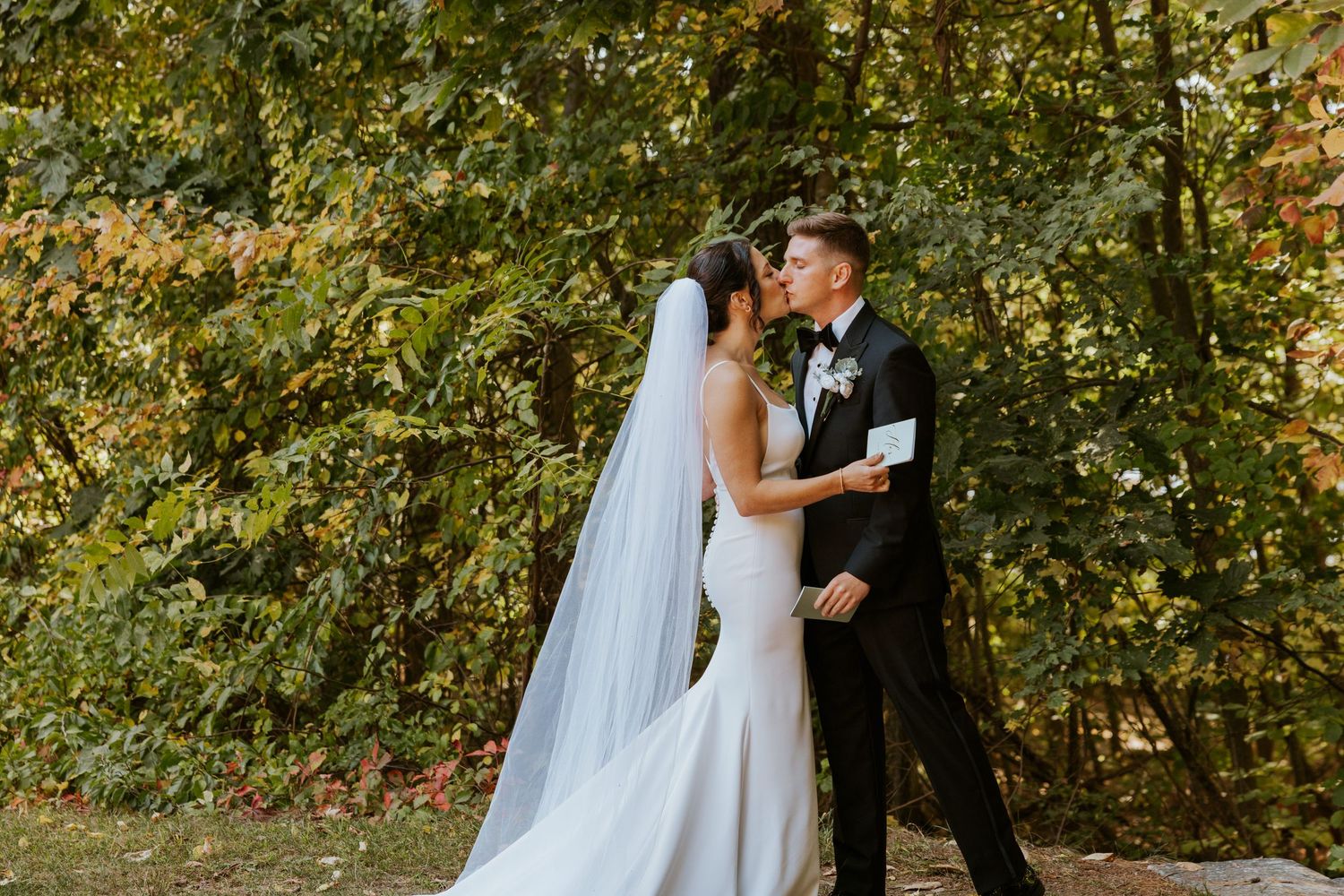 Bride and groom sharing a tender kiss outdoors surrounded by vibrant fall foliage.