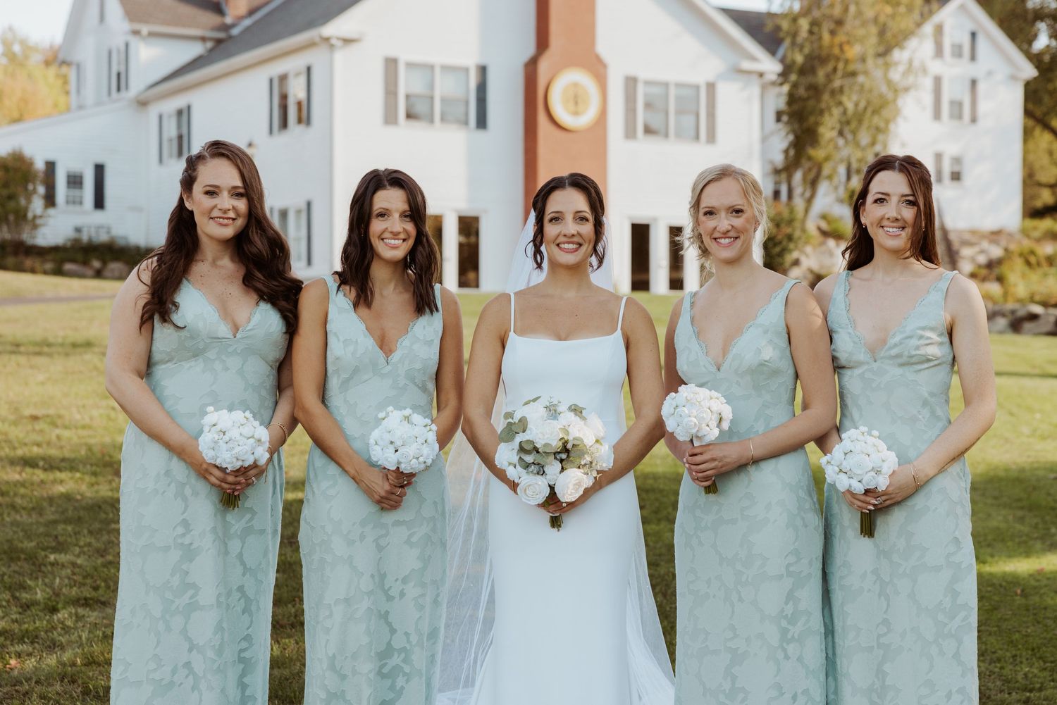 Bride in white gown poses with four bridesmaids in mint lace dresses holding white bouquets outdoors.
