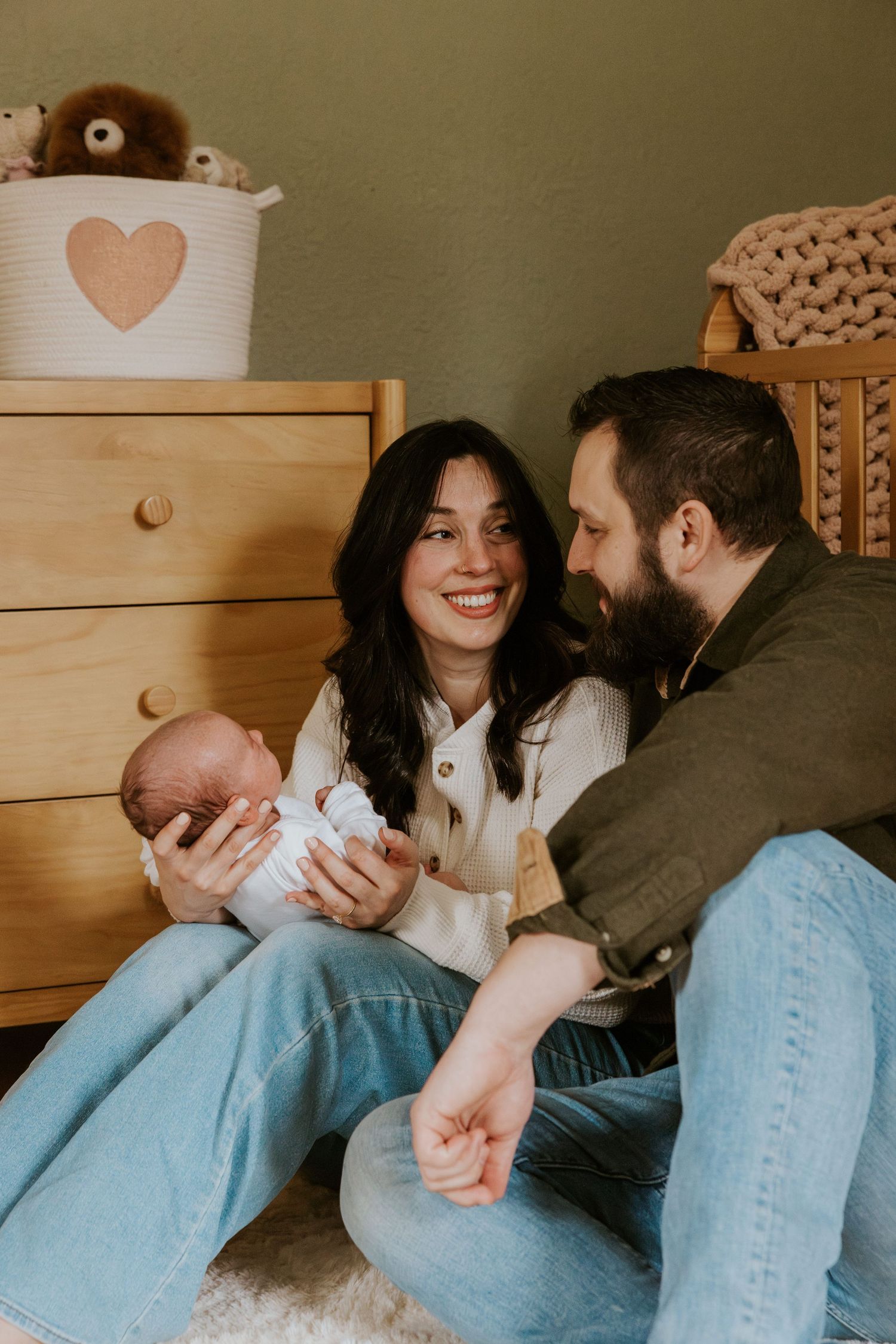 Happy new parents sitting on floor smiling at each other while holding their newborn baby indoors.