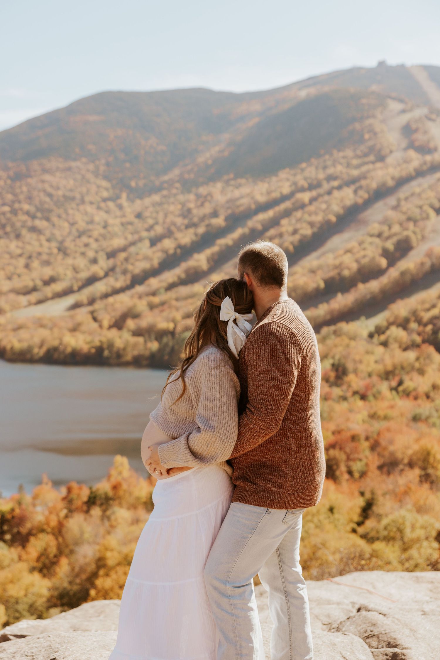Couple embracing on rocky overlook with stunning fall foliage mountain and lake scenery behind them.