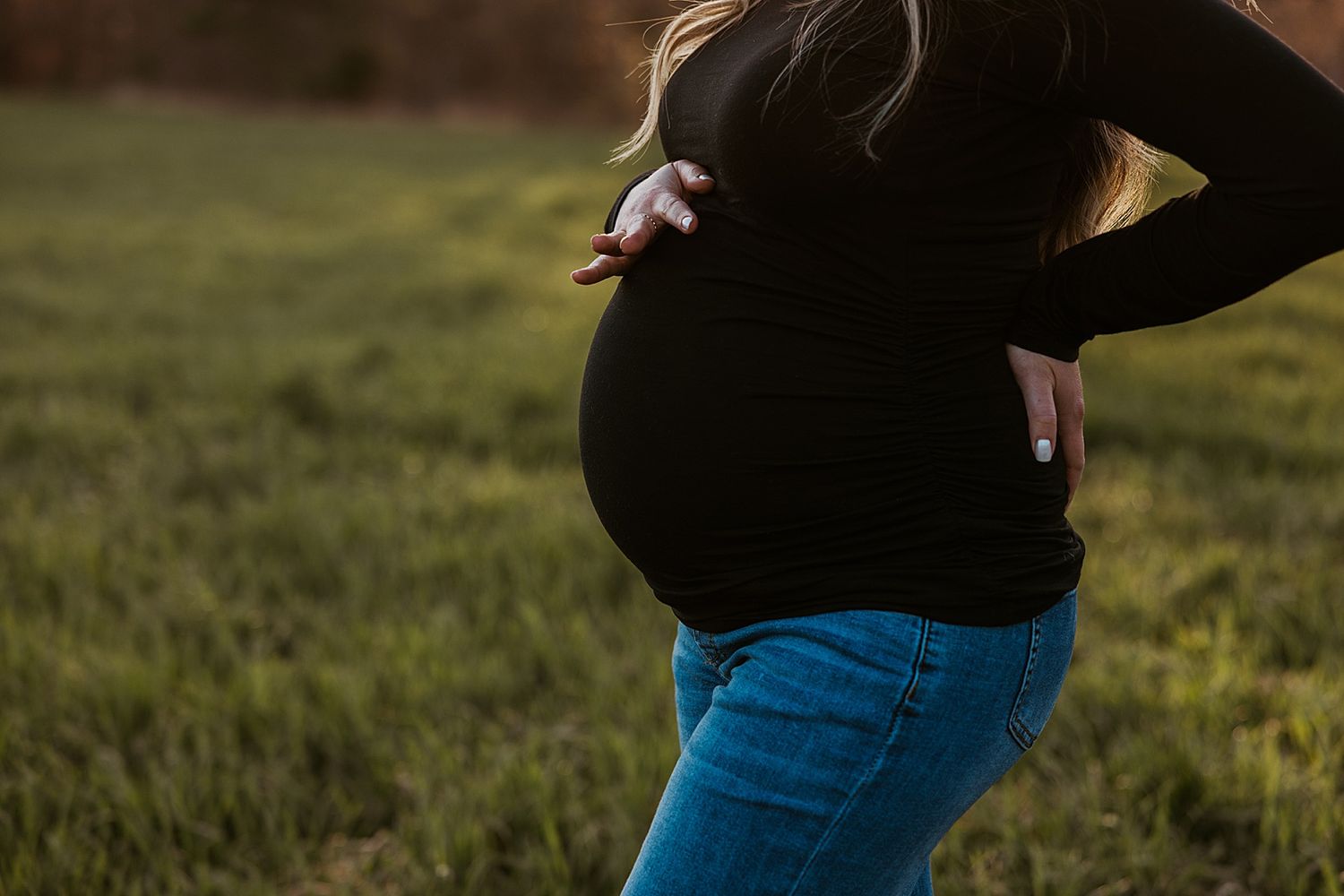 Pregnant woman in black top and jeans cradling baby bump outdoors in fall field during golden hour in New Hampshire