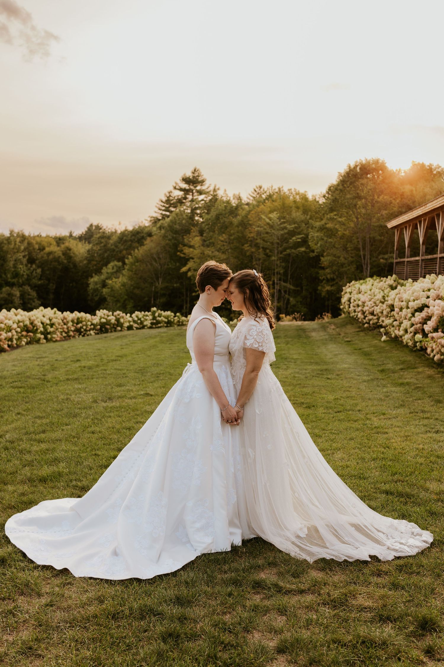 Two brides in flowing white wedding gowns hold hands at sunset on a lush green lawn lined with white flowers.