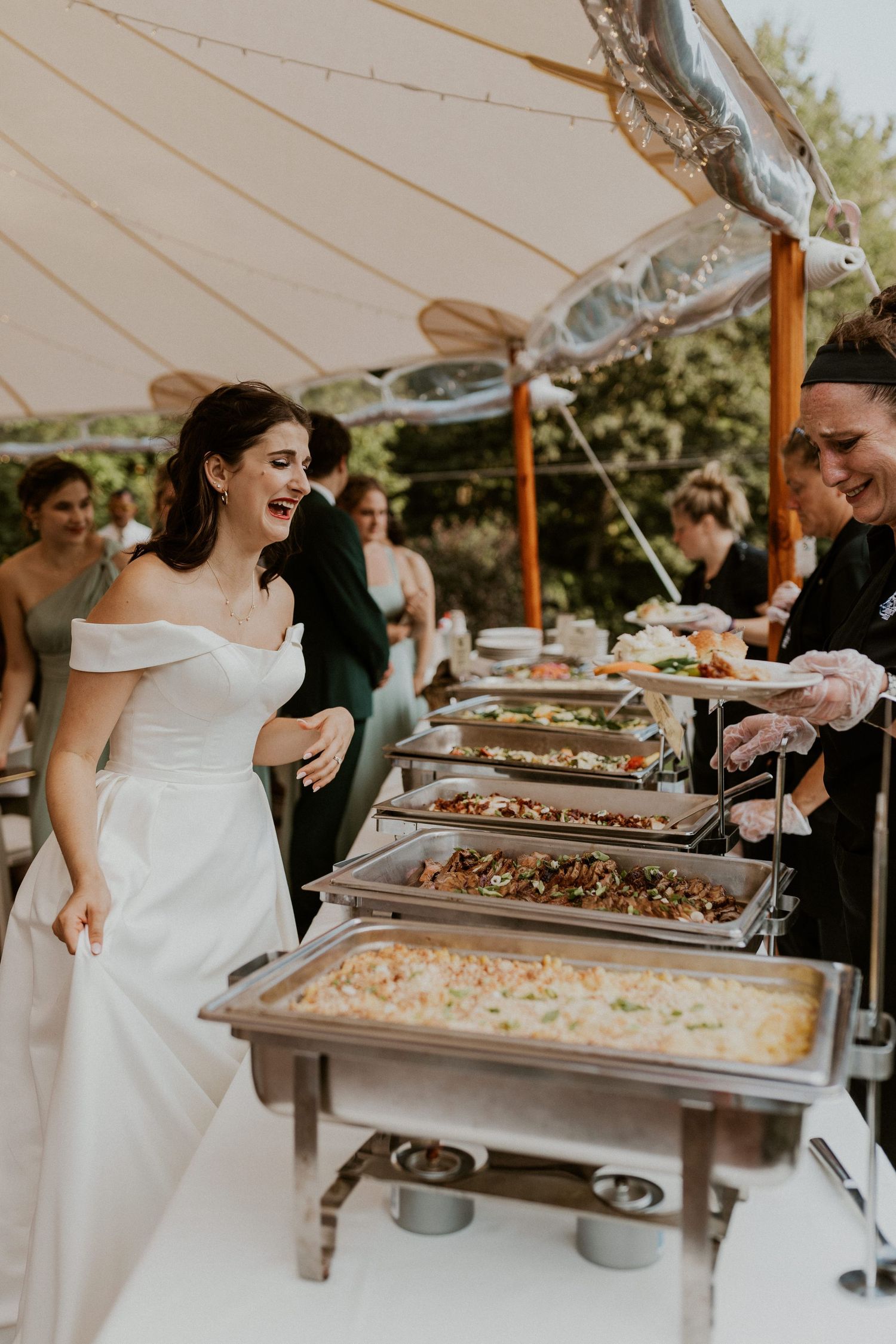 Elegant buffet line with chafing dishes under white tent at outdoor wedding reception.