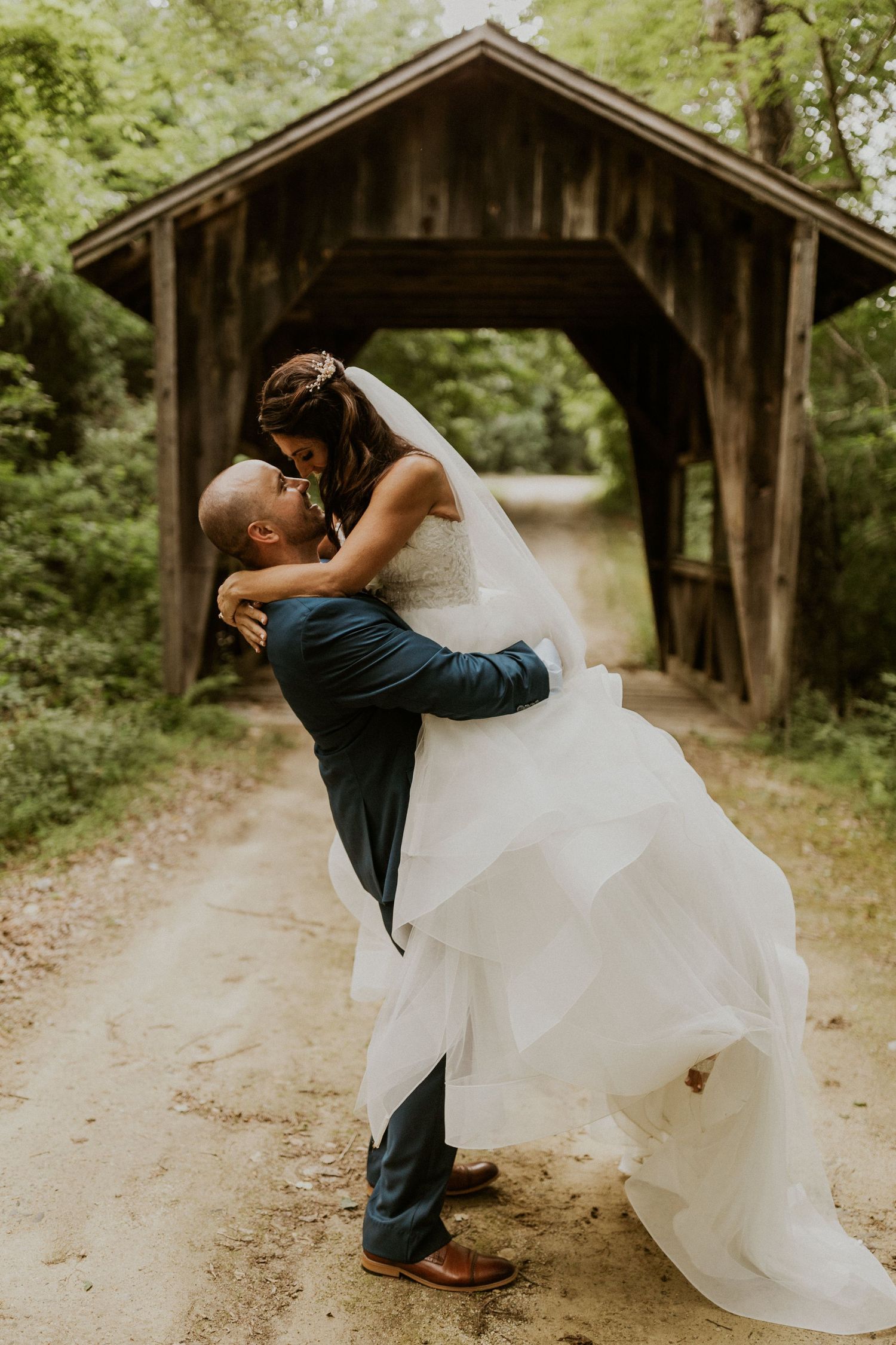 Romantic wedding couple poses near wooden covered bridge in forest setting.
