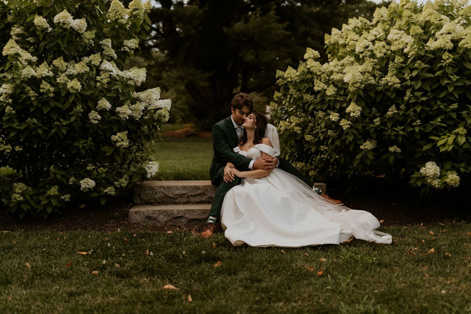 A romantic wedding couple sitting on garden steps surrounded by blooming white hydrangeas.