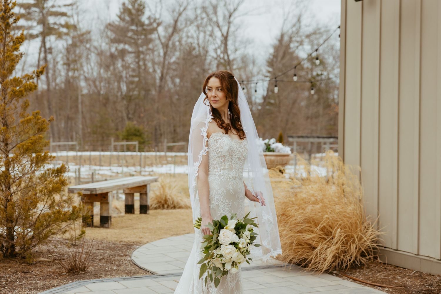 Bride in white dress holding bouquet while standing in front of modern building on autumn day.