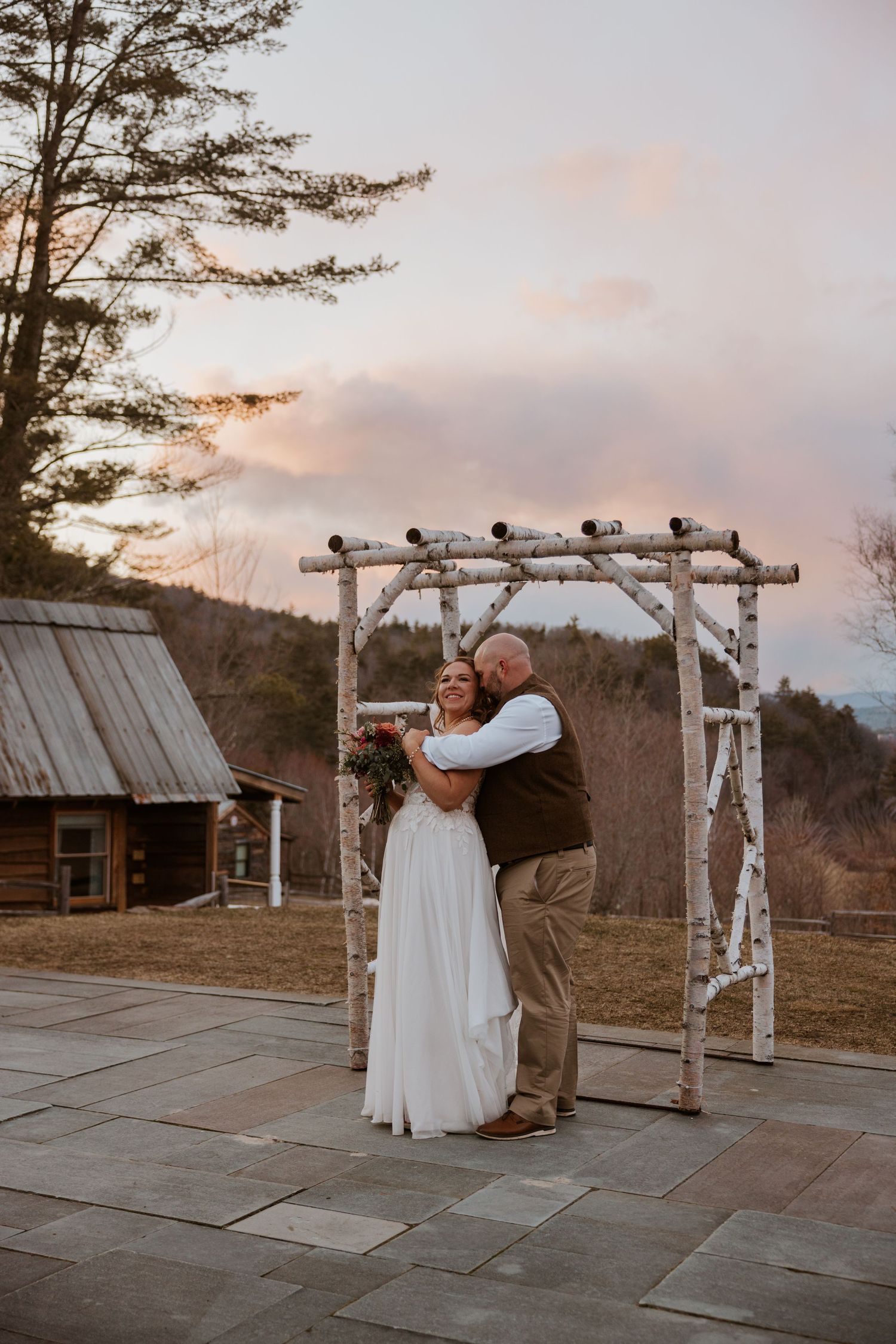 A romantic sunset wedding moment under a birch arch on a stone patio.